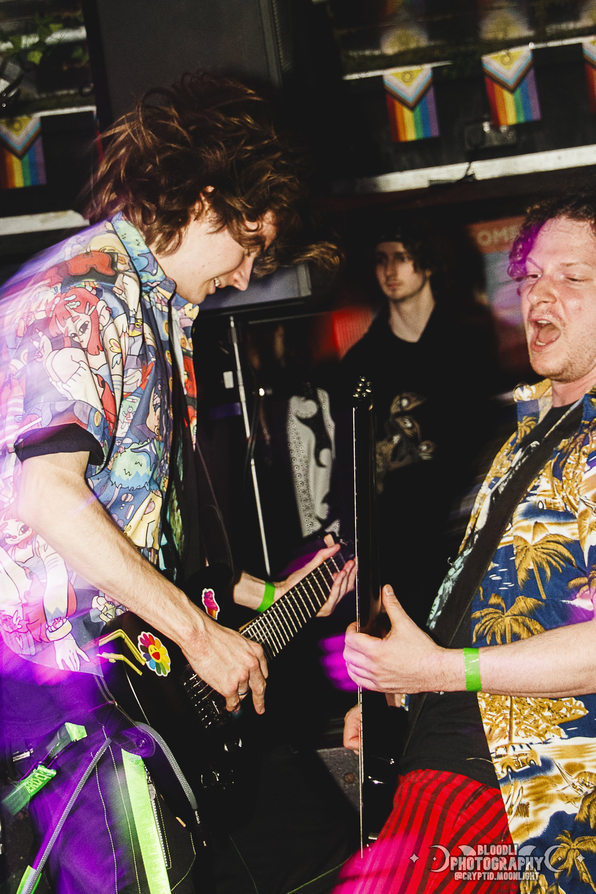 Two individuals playing guitars and singing passionately on stage at a lively concert, wearing colorful Hawaiian shirts, with a person in the background watching.