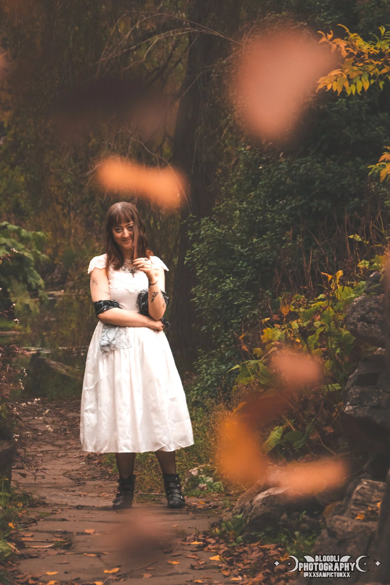 A woman standing on a wooded path surrounded by trees and rocks with orange leaves falling around her.