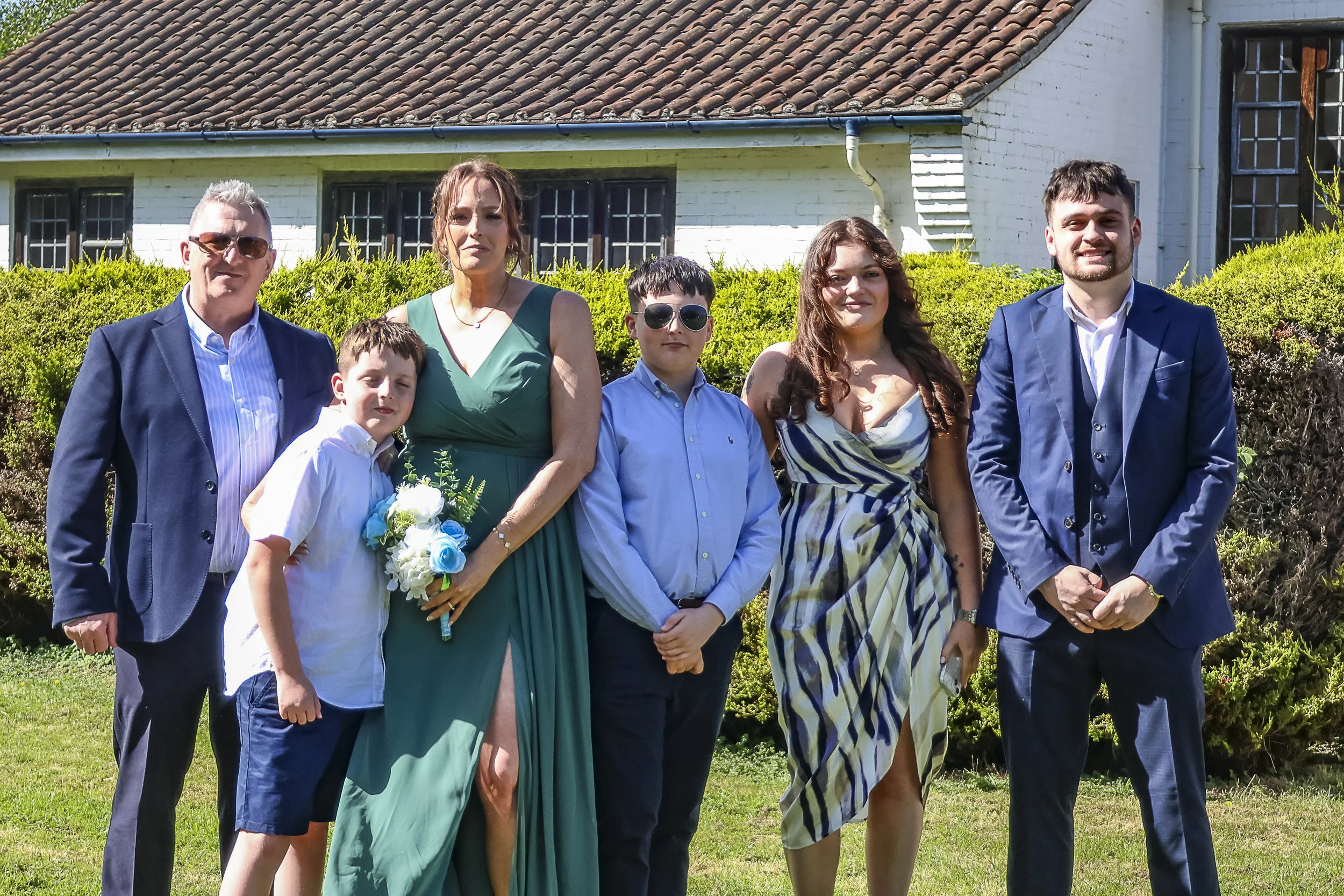 Family group photo outdoors in front of a house with white brick and a tiled roof, featuring two adults and four children, with a backdrop of green bushes.