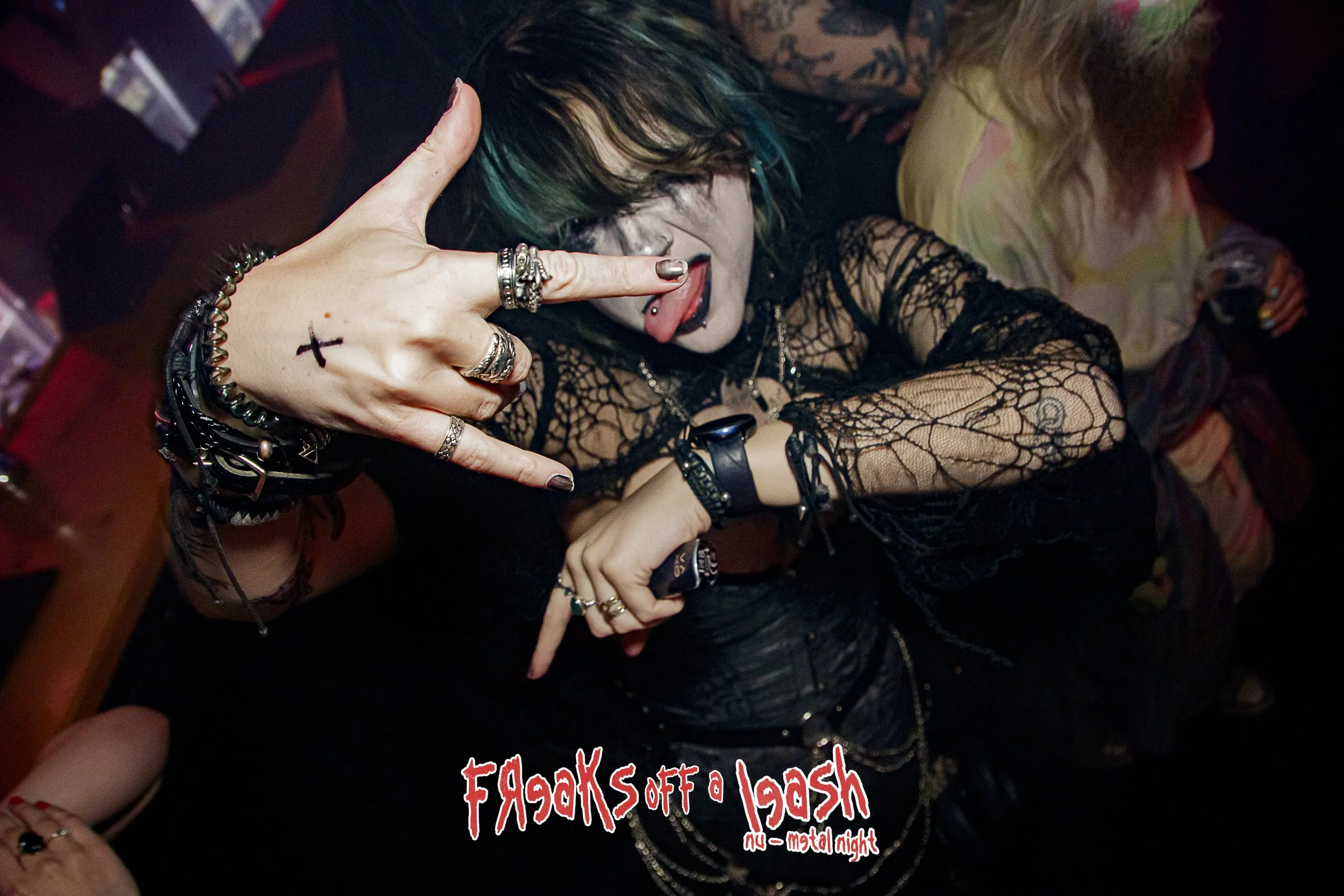 A person with dark hair and blue hair highlights sticking their tongue out and making a rock and roll hand gesture with their fingers. They are wearing multiple silver rings and bracelets, and a black lace top. The background shows a person with blonde hair and a multicolored shirt. There is red and white text at the bottom of the image that says "FREAKS off a lash" and "metal night."