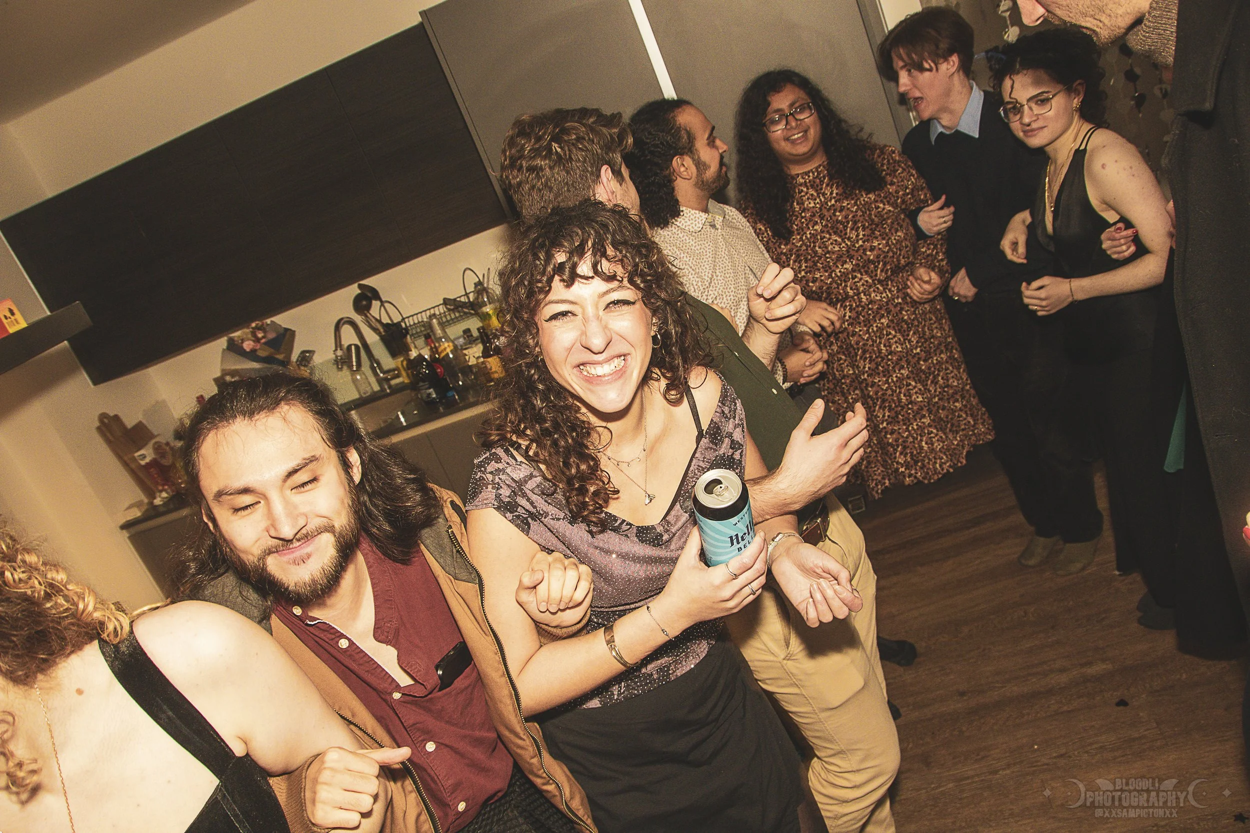 Group of people at a party, smiling, socializing, and having fun in a cozy room with a kitchen in the background.