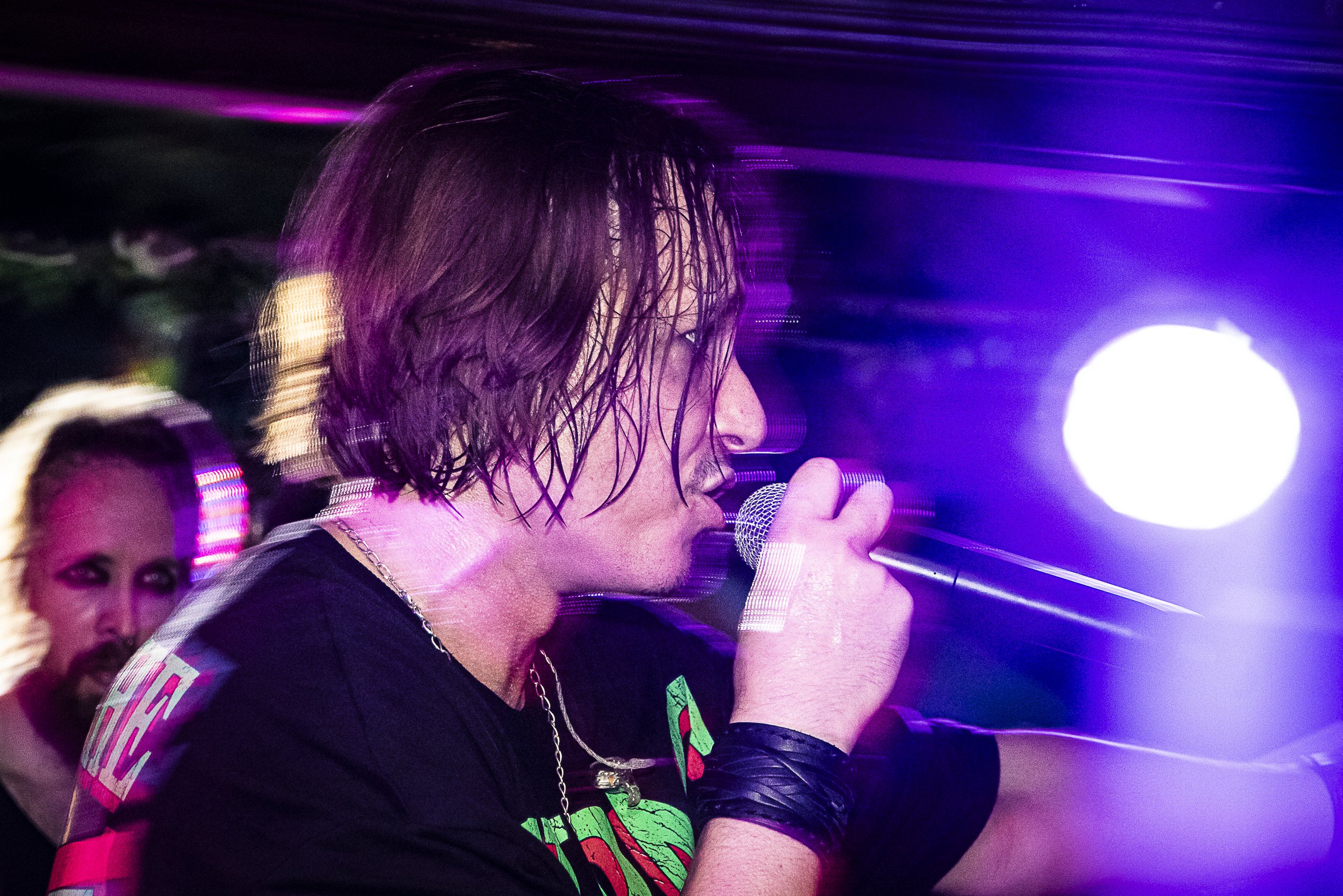A male singer with shoulder-length hair sings into a microphone on stage, illuminated by purple and white lights, with a woman with blonde hair and dark eye makeup visible in the background.