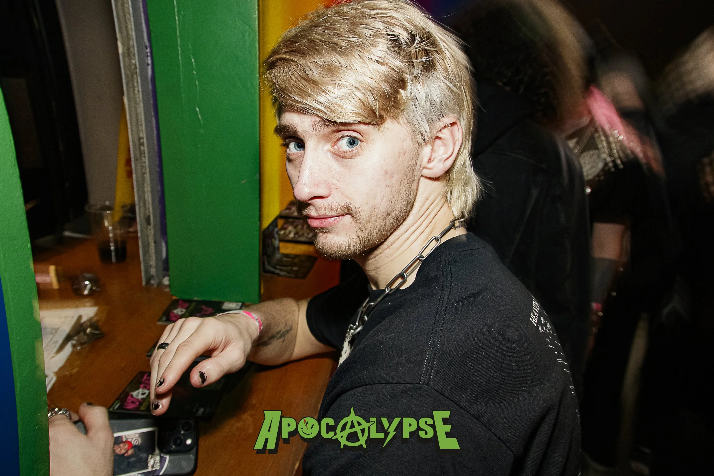 A young man with blond hair and blue eyes sitting at a gaming machine in a nightclub or arcade, wearing a black shirt and a statement chain necklace, with the logo 'APOCALYPSE' at the bottom of the image.