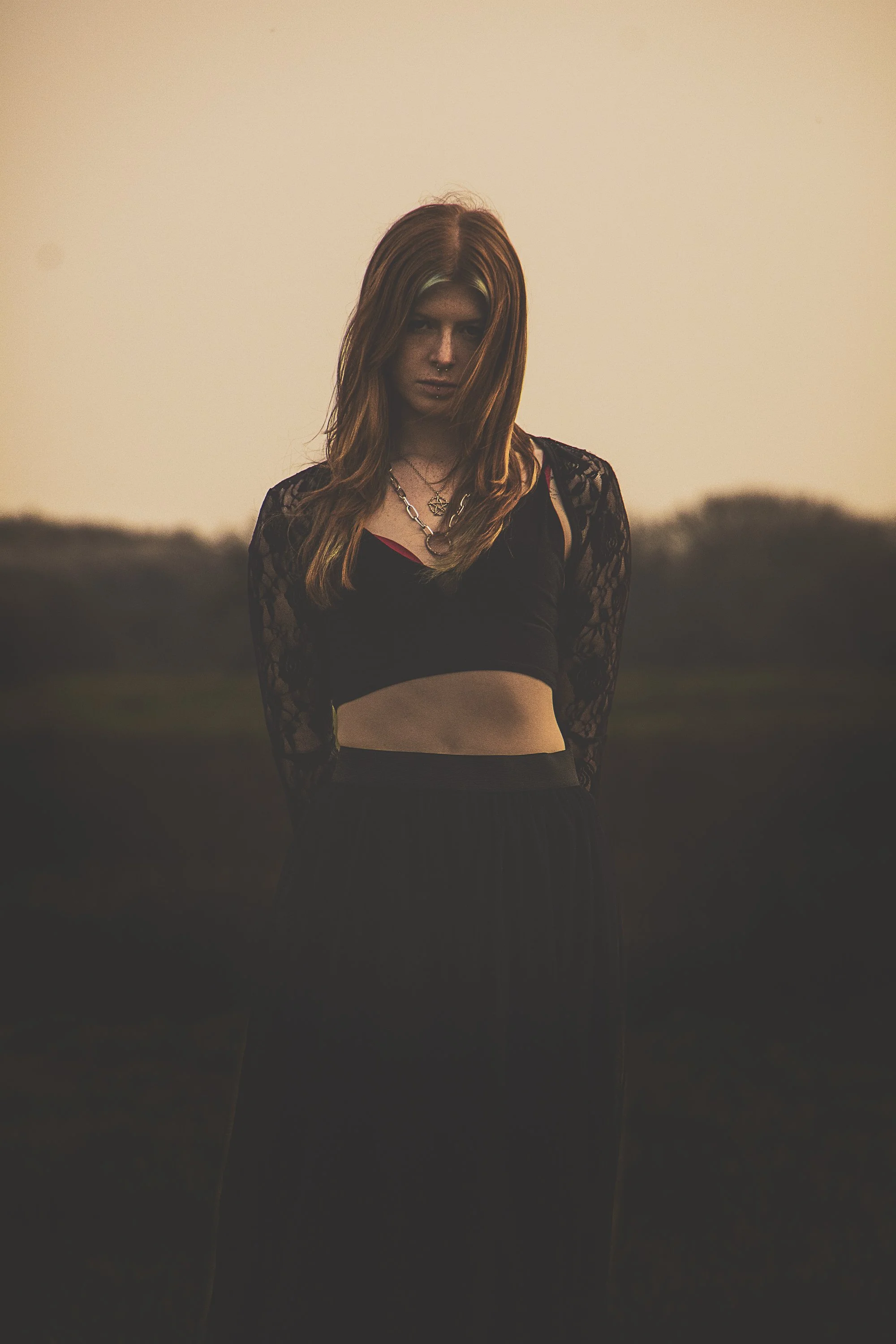 Young woman with long red hair wearing a black crop top with lace sleeves and a black skirt, standing outdoors during sunset with a blurred natural background.