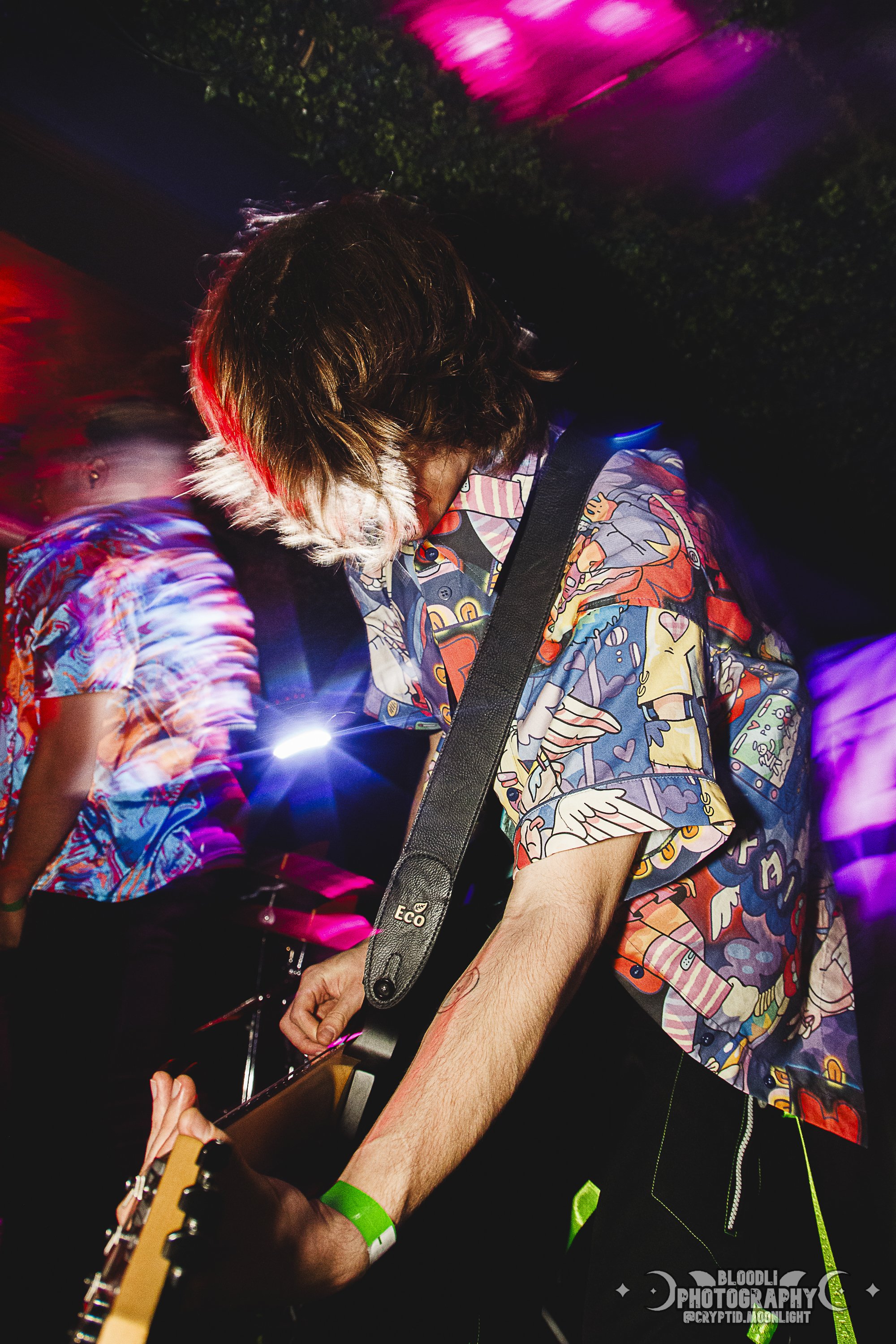 Person playing guitar at a concert, wearing a colorful, patterned shirt and a green wristband, with vibrant pink, purple, and red lights in the background.