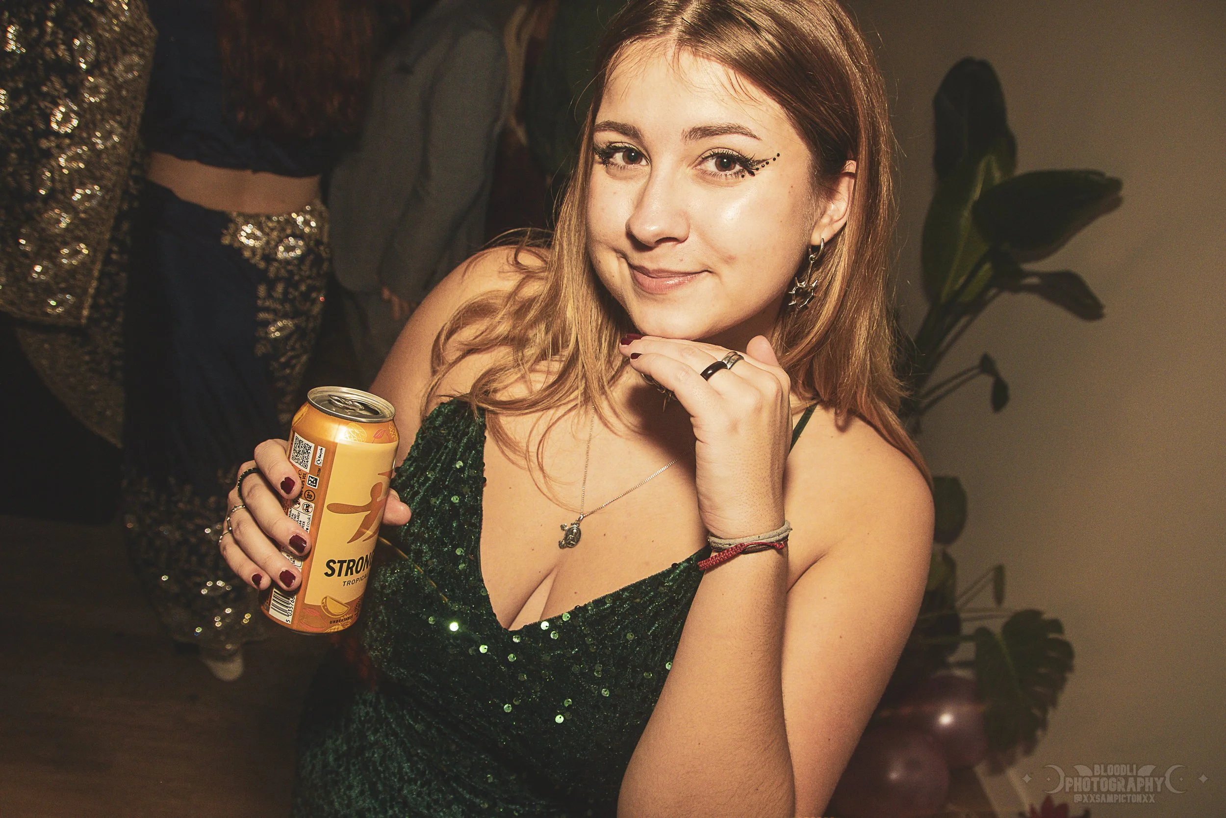 A young woman with light brown hair wearing a green sequin dress, accessorized with earrings and necklaces, smiling at the camera while holding a can of Strong Tropical drink at a party.