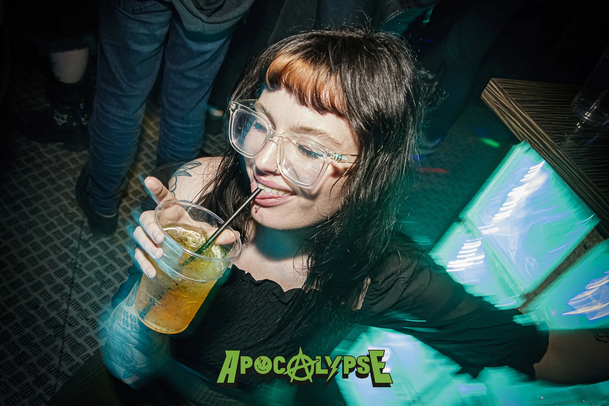 A young woman with black hair, glasses, and piercings, wearing a black top, is drinking a drink with a straw at a party or club. There are other people in the background and colorful lighting.