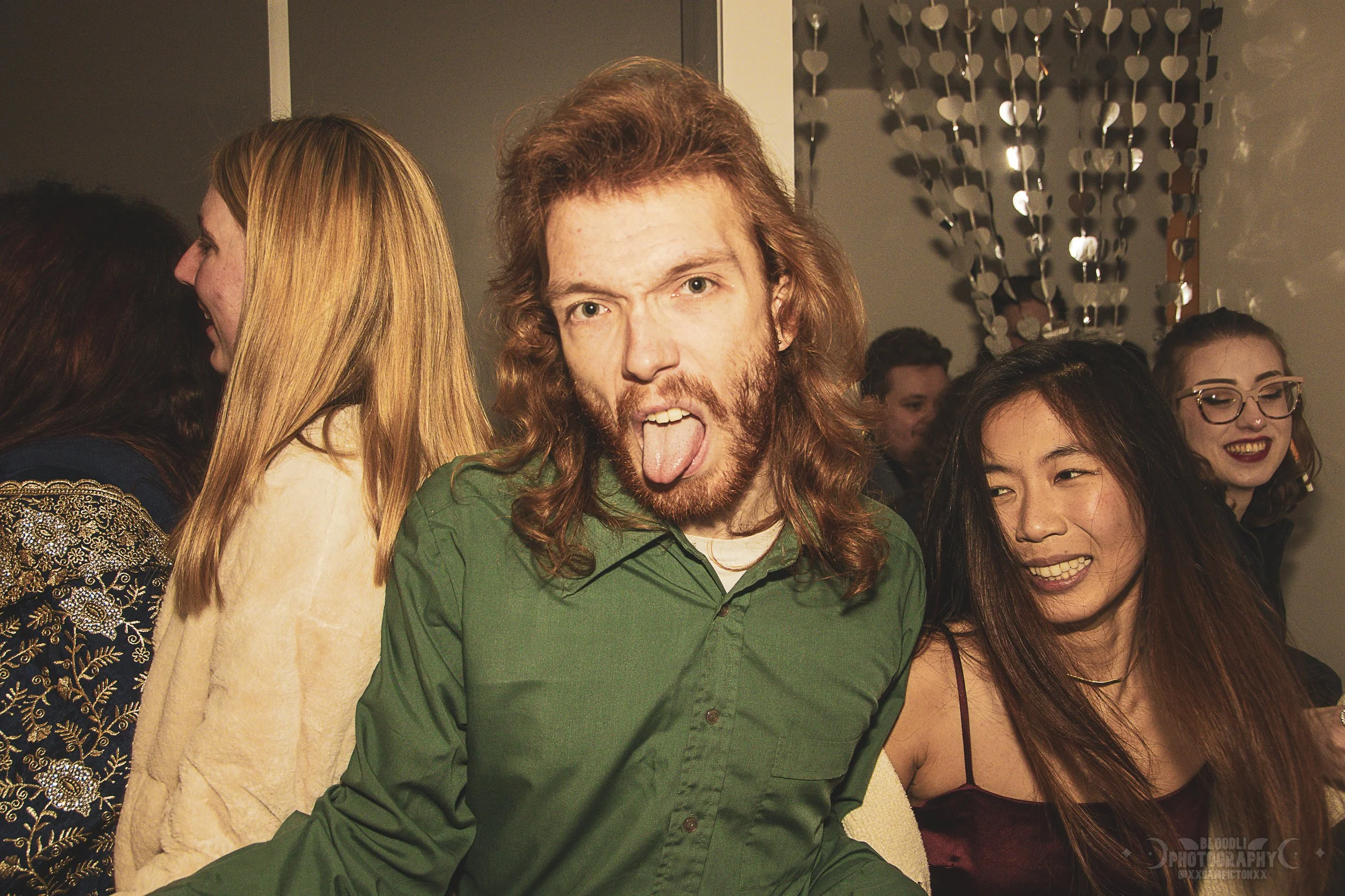 A group of young people at a social gathering or party, with one man in the foreground sticking out his tongue and posing playfully. There are women beside and behind him, all smiling or laughing. The background shows a wall decorated with hanging me