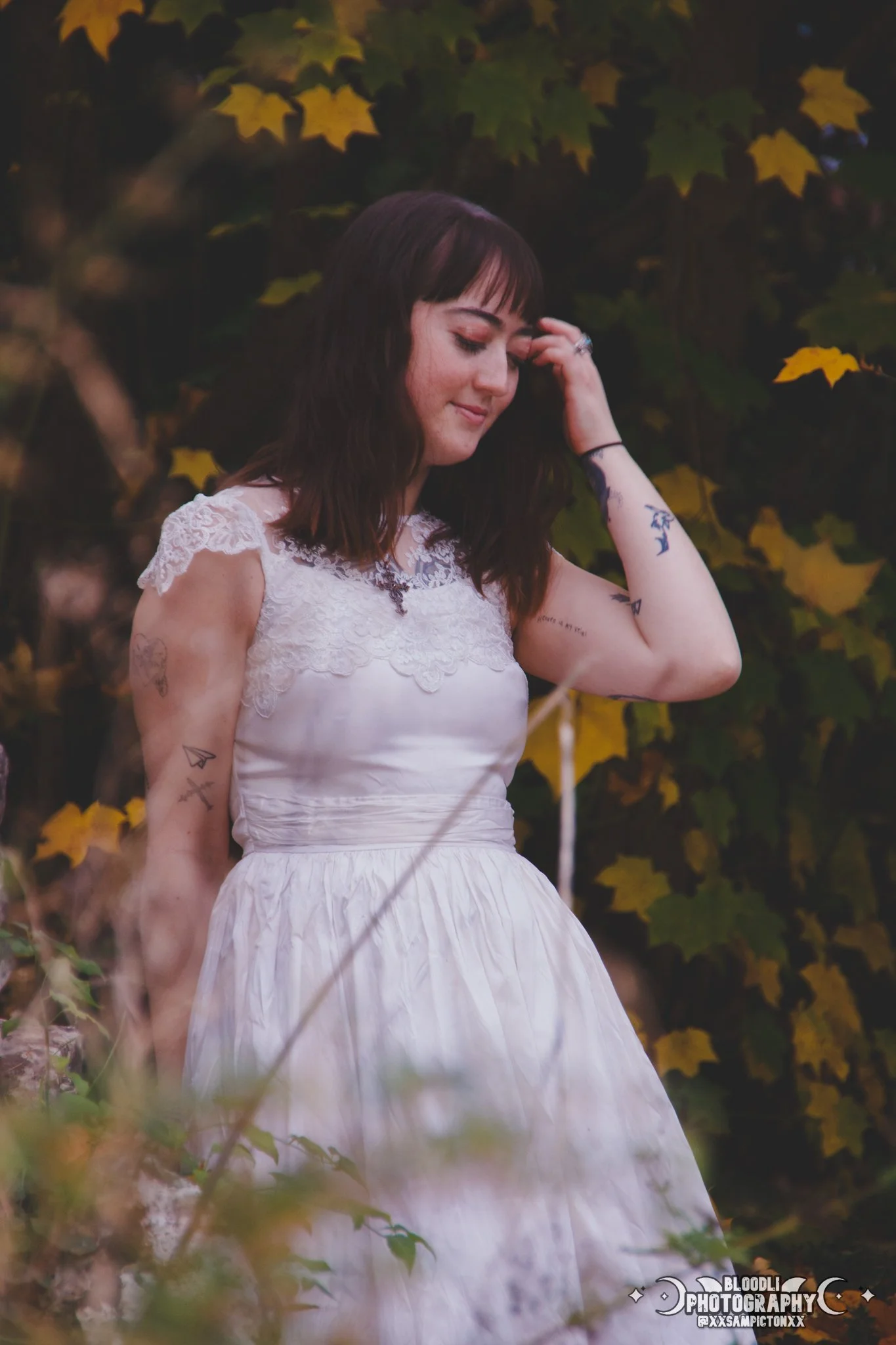 A woman with dark brown hair and tattoos, wearing a white lace dress, standing outdoors against a background of green and yellow leaves, with her eyes closed and hand touching her hair.