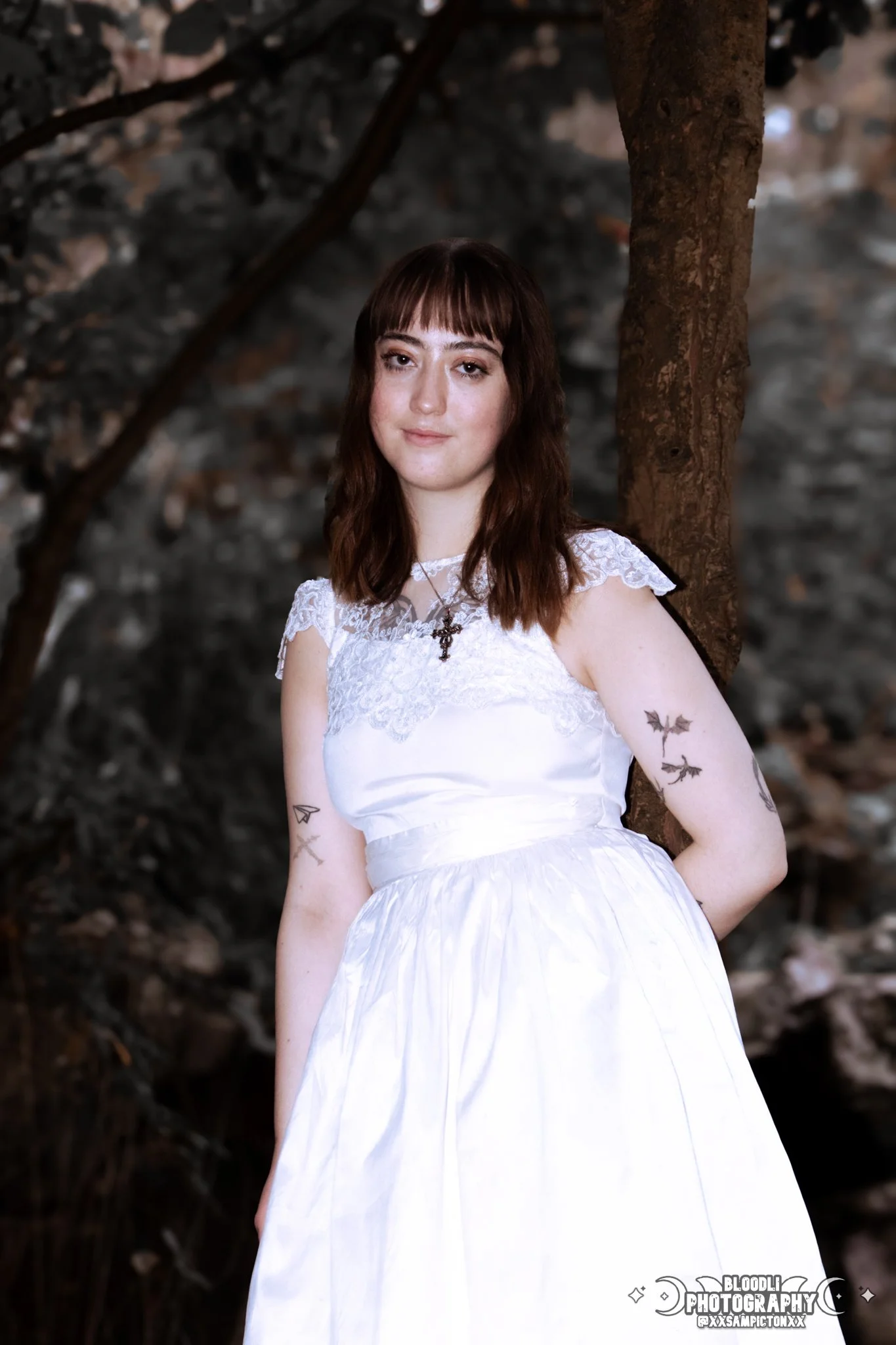 A young woman in a white dress standing outdoors in front of a tree and a dark background.