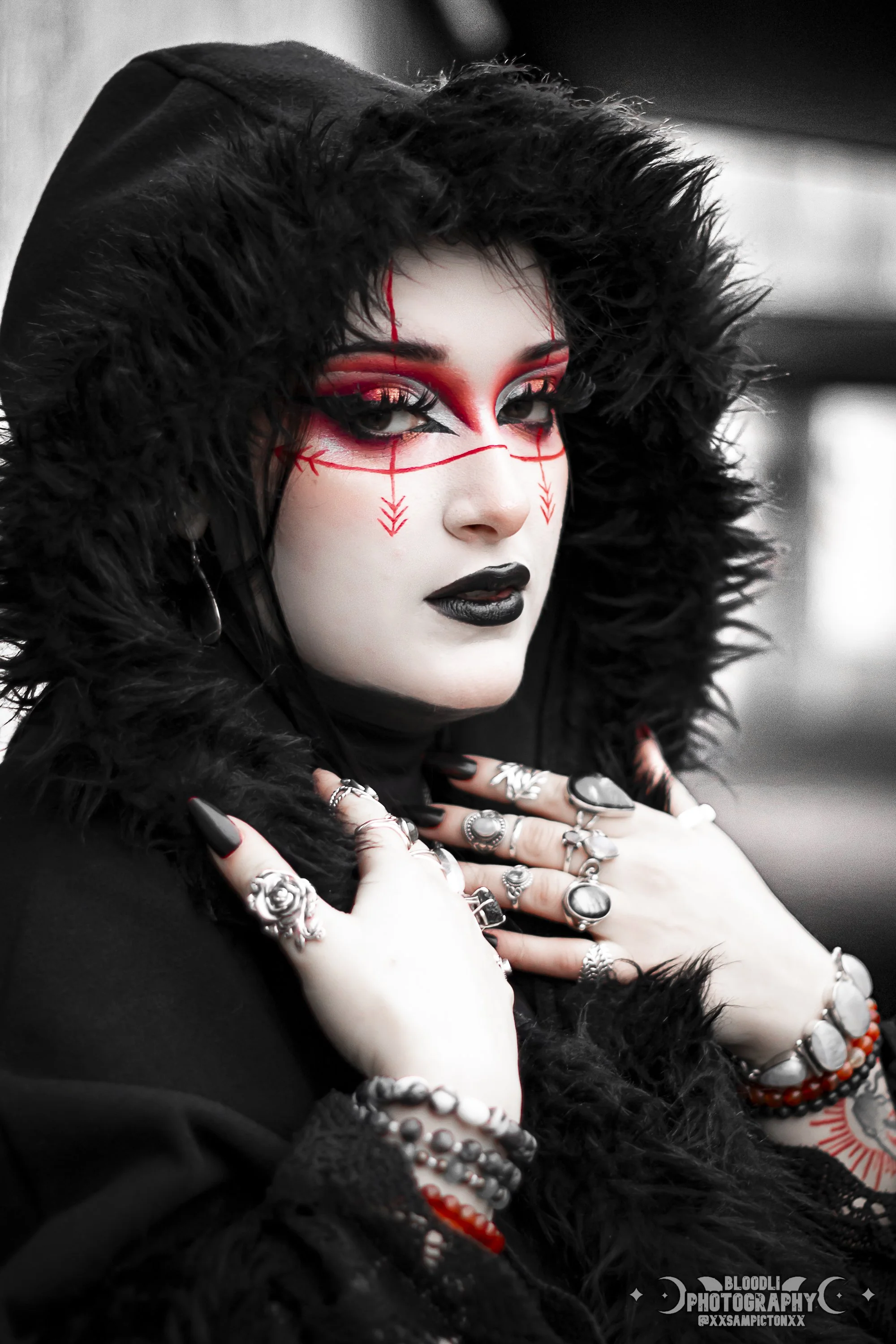 A woman with dramatic makeup and jewelry, wearing a black hood with fur trim. Her makeup includes bold black and red eye makeup, black lipstick, and red decorative lines on her face. She has multiple rings and bracelets and poses with her hands near her face, showing her rings and nails.