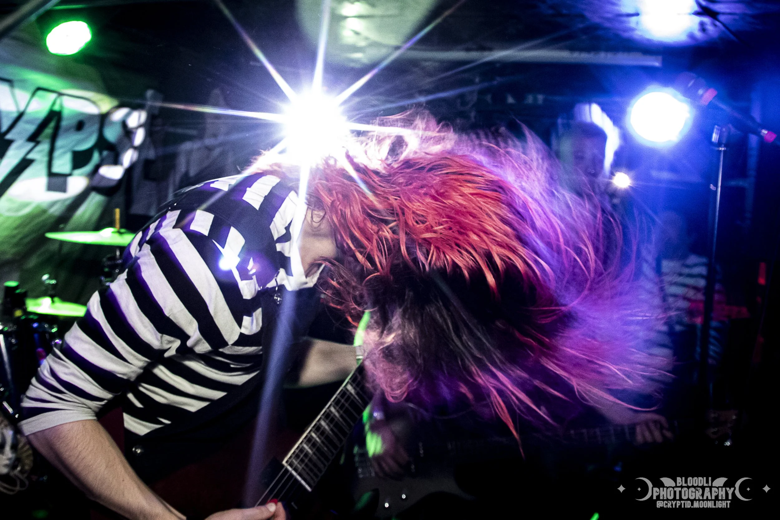 Person with long red hair playing guitar on stage in a dimly lit venue with colorful stage lights and a neon sign in the background.