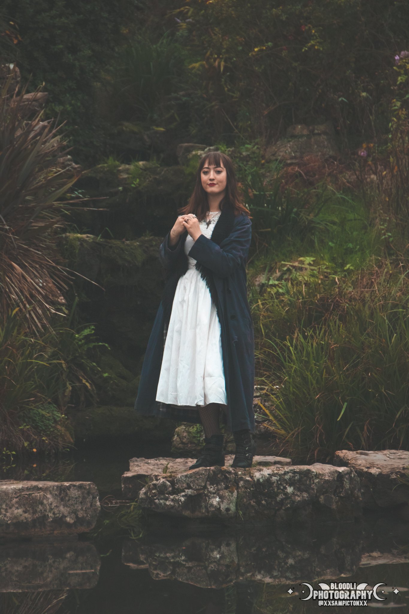 A woman dressed in a historical or vintage style white dress with a dark coat standing on rocks by a water stream in a lush, green outdoor setting.