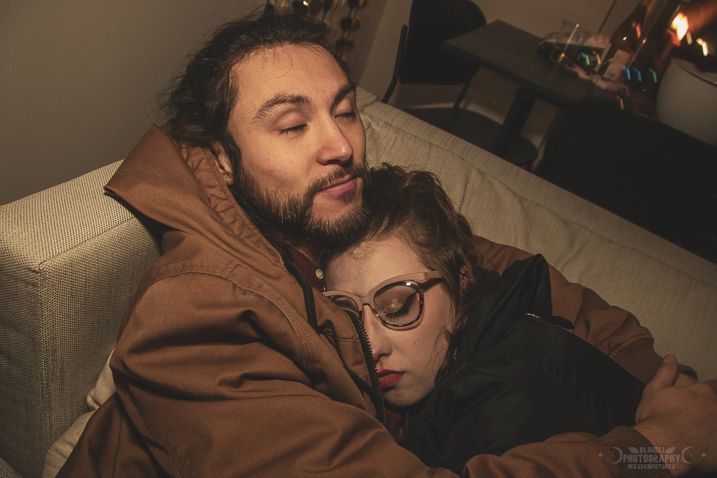 A man with a beard and long hair, wearing a brown jacket, is sitting on a beige couch with a woman with curly hair and glasses, wearing a black jacket, who is sleeping with her head on his chest, both appearing peaceful and content.