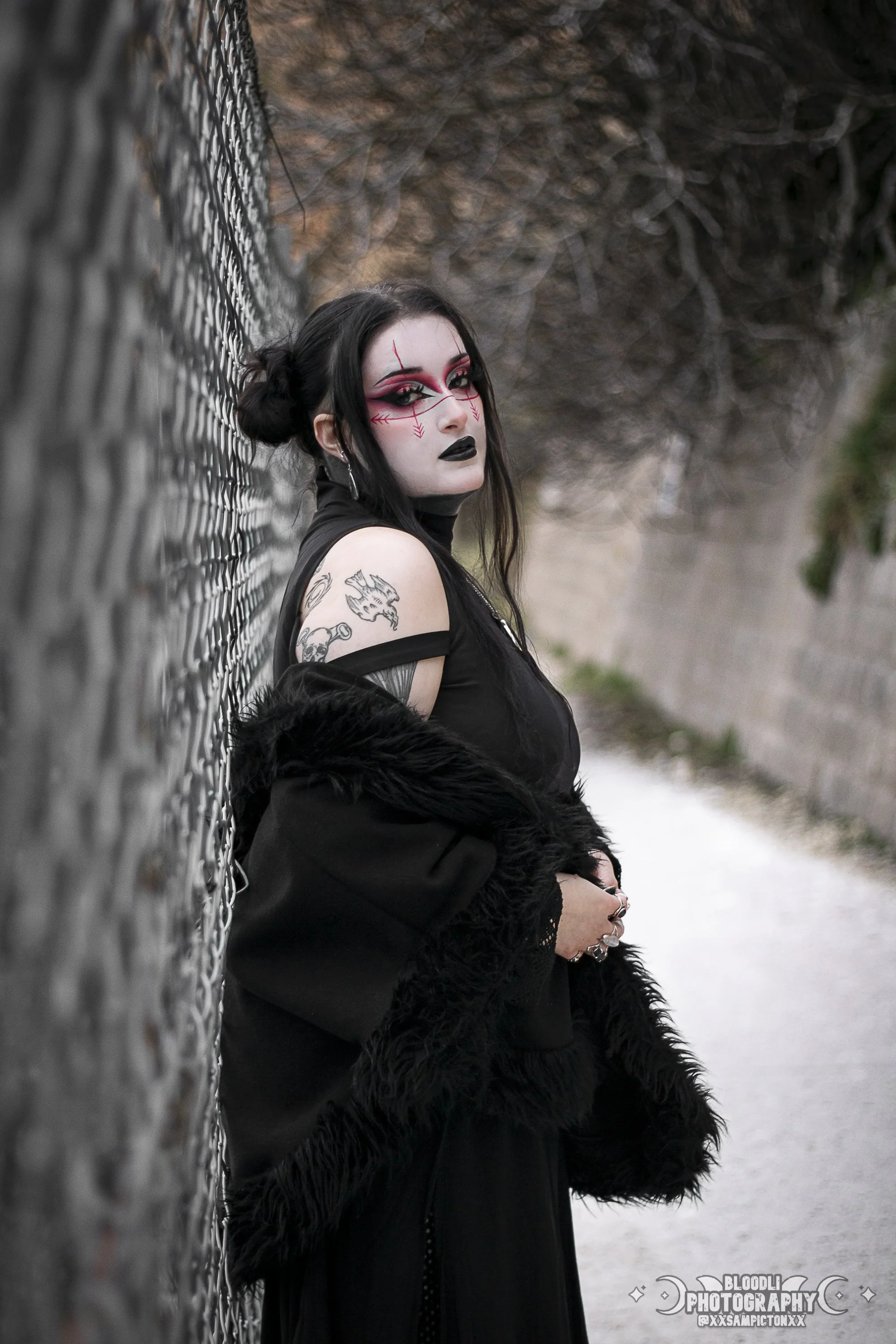 A woman with gothic makeup and tattoos standing outdoors against a chain-link fence, with a blurred natural background.