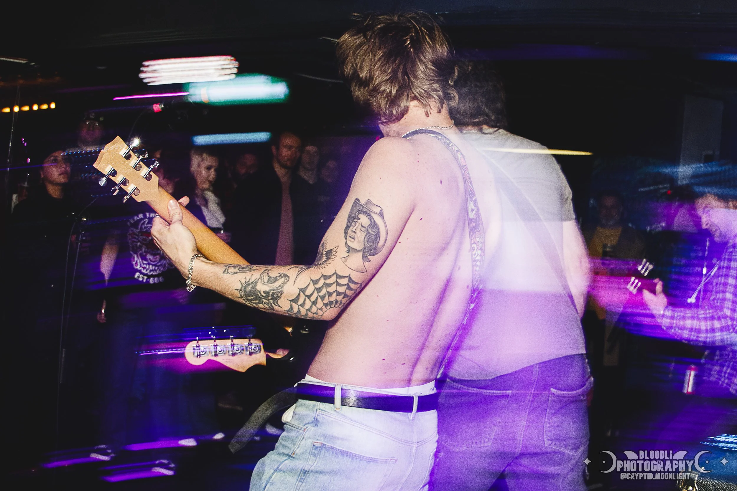 T-shirtless guitarist playing electric guitar at a dimly lit concert with audience in the background, motion blur and colorful lights.