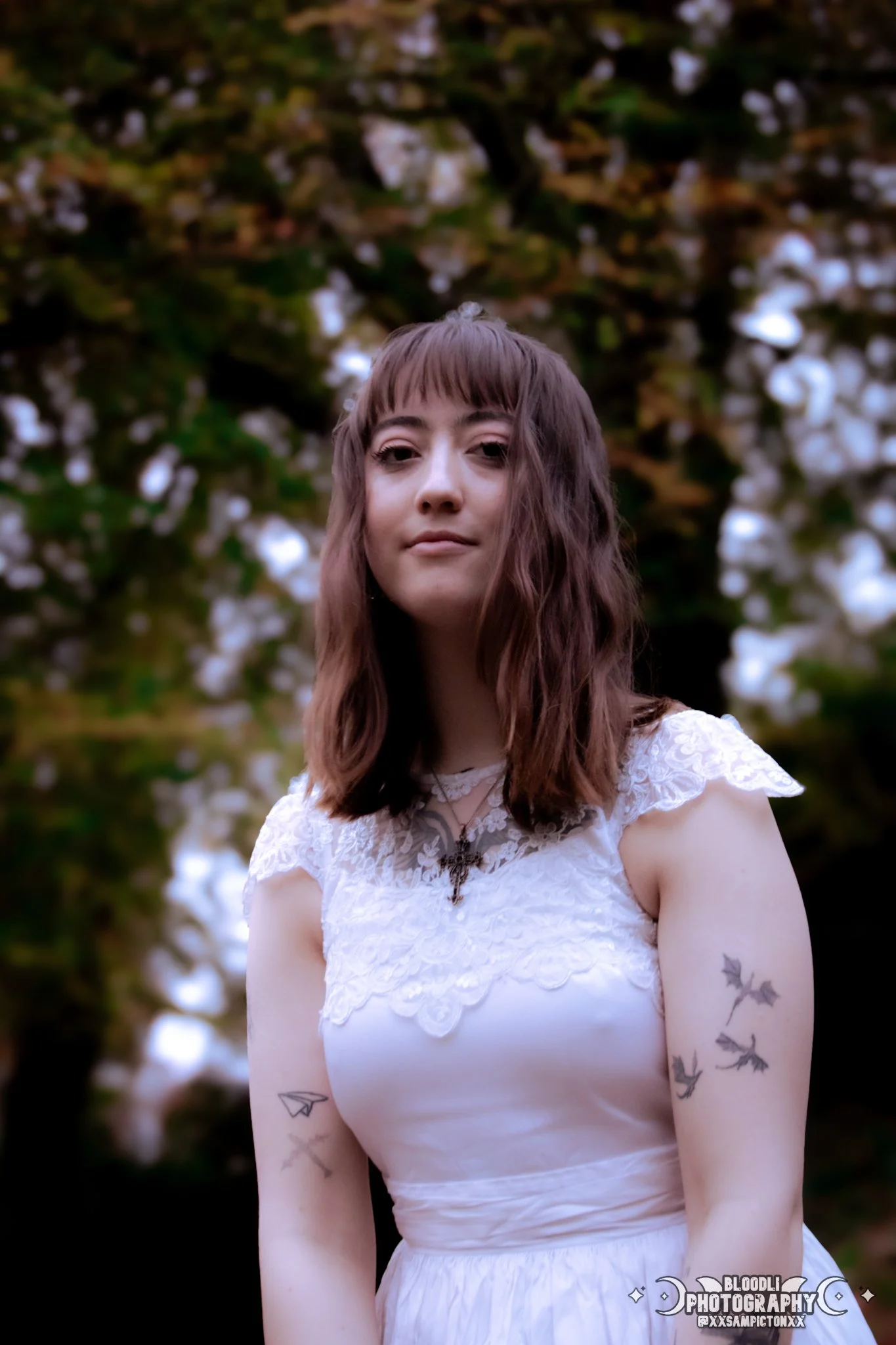 A young woman with shoulder-length brown hair and bangs, wearing a white lace dress with short sleeves, stands outdoors in front of a background of trees with green leaves and white flowers. She has tattoos of flying birds on her right arm and a pape