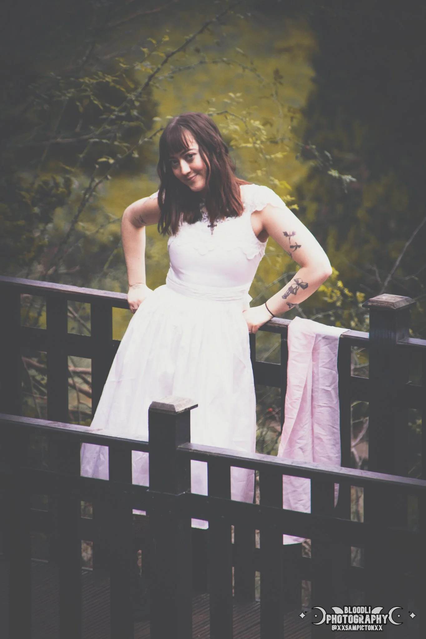 A woman with tattoos on her arms, wearing a white dress, standing on a wooden deck with a forest background, leaning on the railing with her hands on her hips, smiling at the camera.