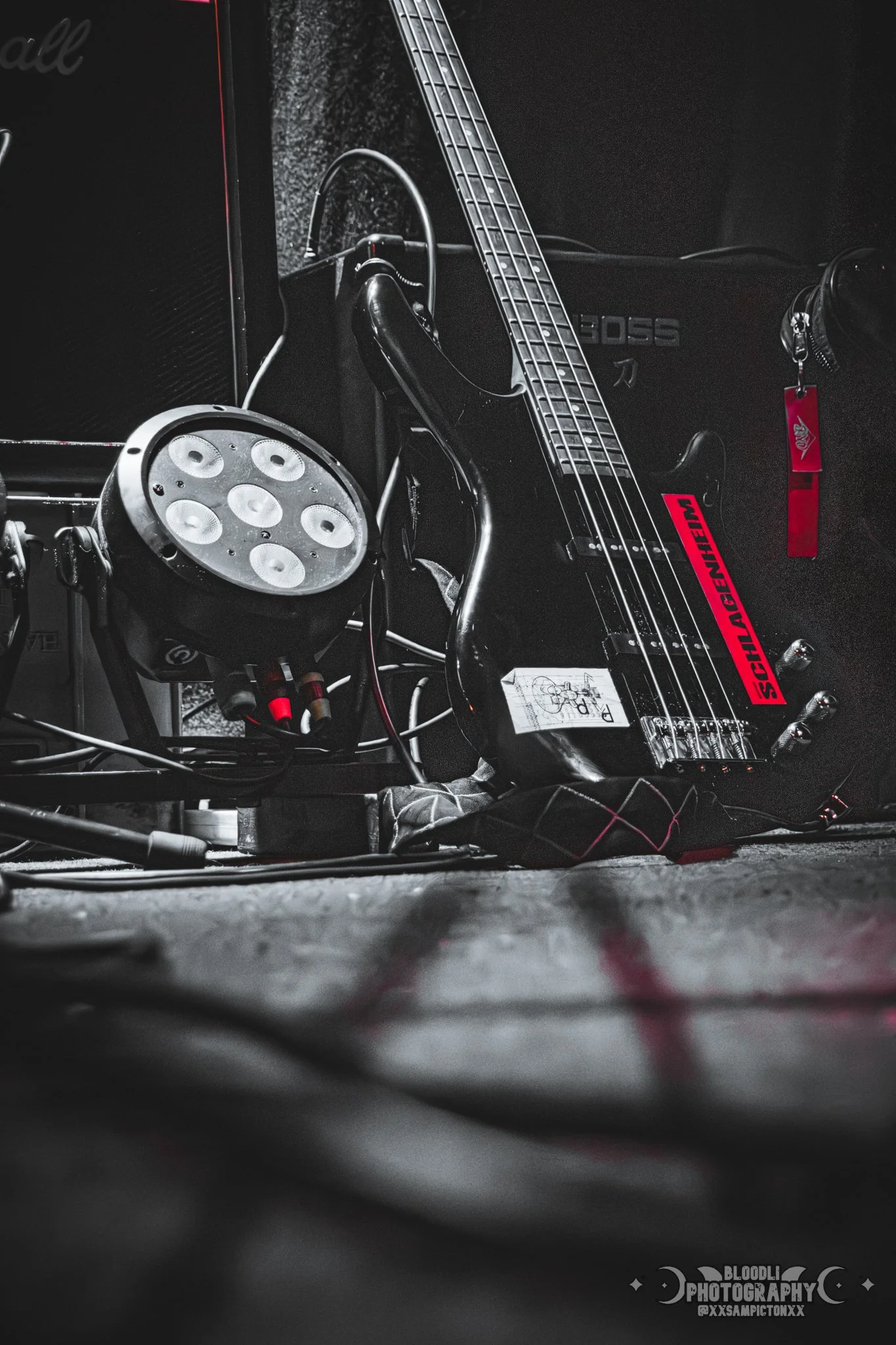 A black electric guitar with red stickers leaning against a black amplifier, next to a black lighting fixture on a dark floor.