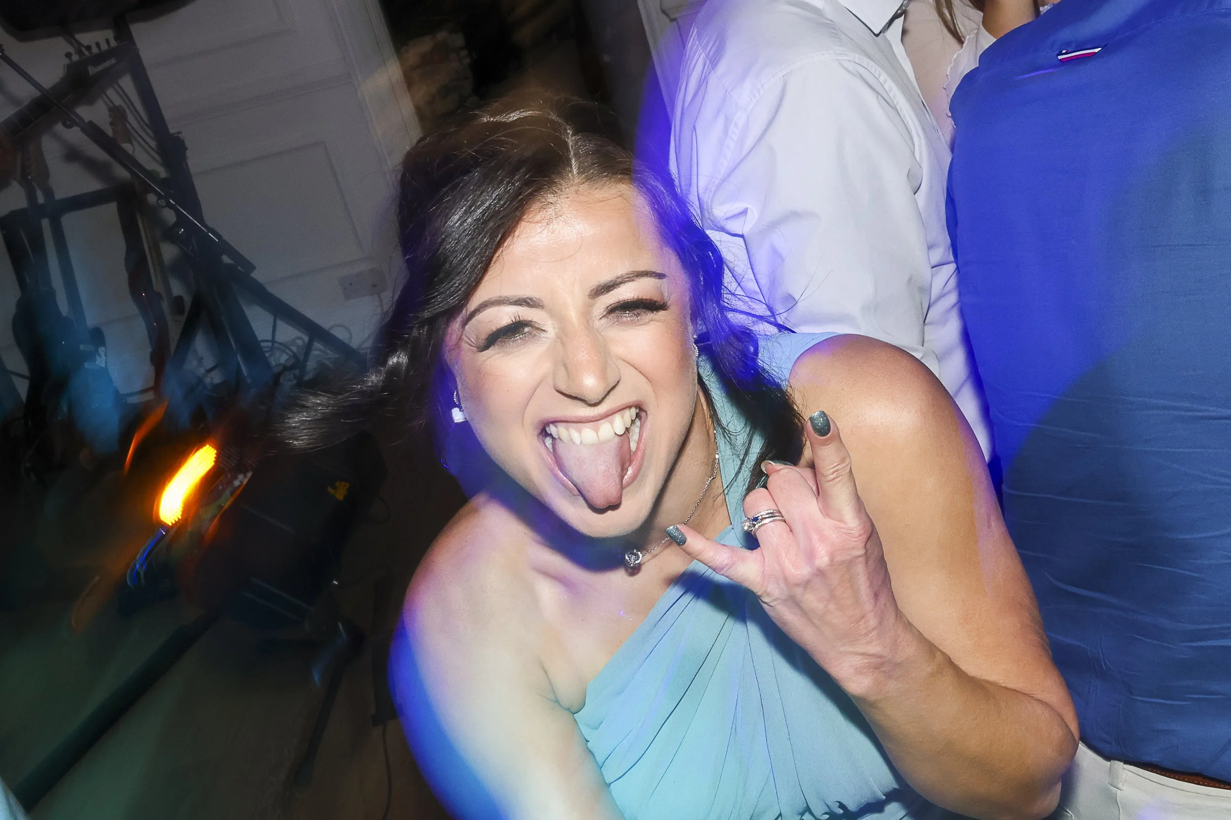A woman with dark hair smiling and sticking out her tongue at a party, showing her hand with rings, wearing a teal dress.