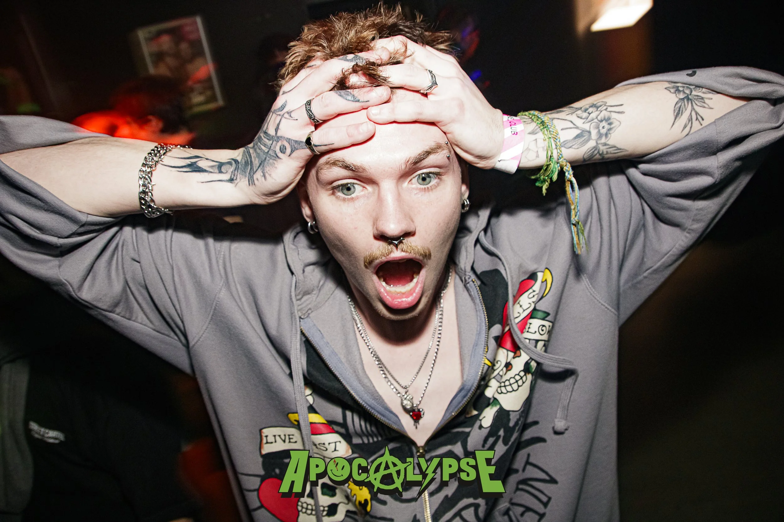 Person with short red hair and tattoos on arms, wearing a gray hoodie with a graphic skull design, holding head with hands, with mouth open in surprise or shock, at a party.