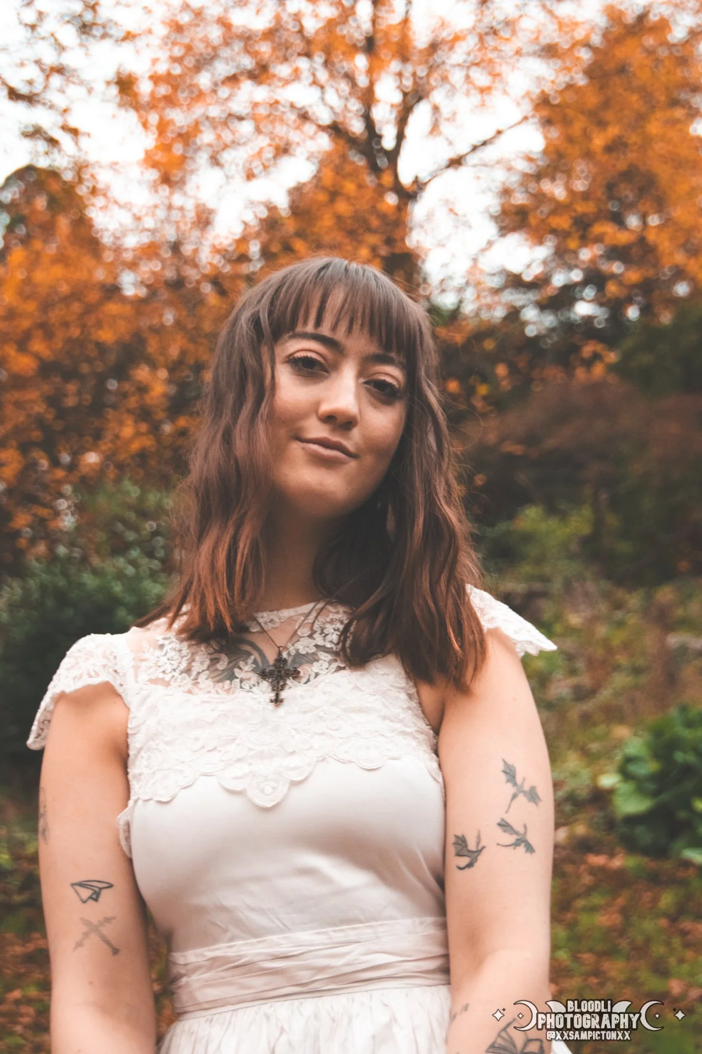 Young woman with brown hair and tattoos on her arms, wearing a cream-colored lace dress, standing outdoors with autumn trees in the background.