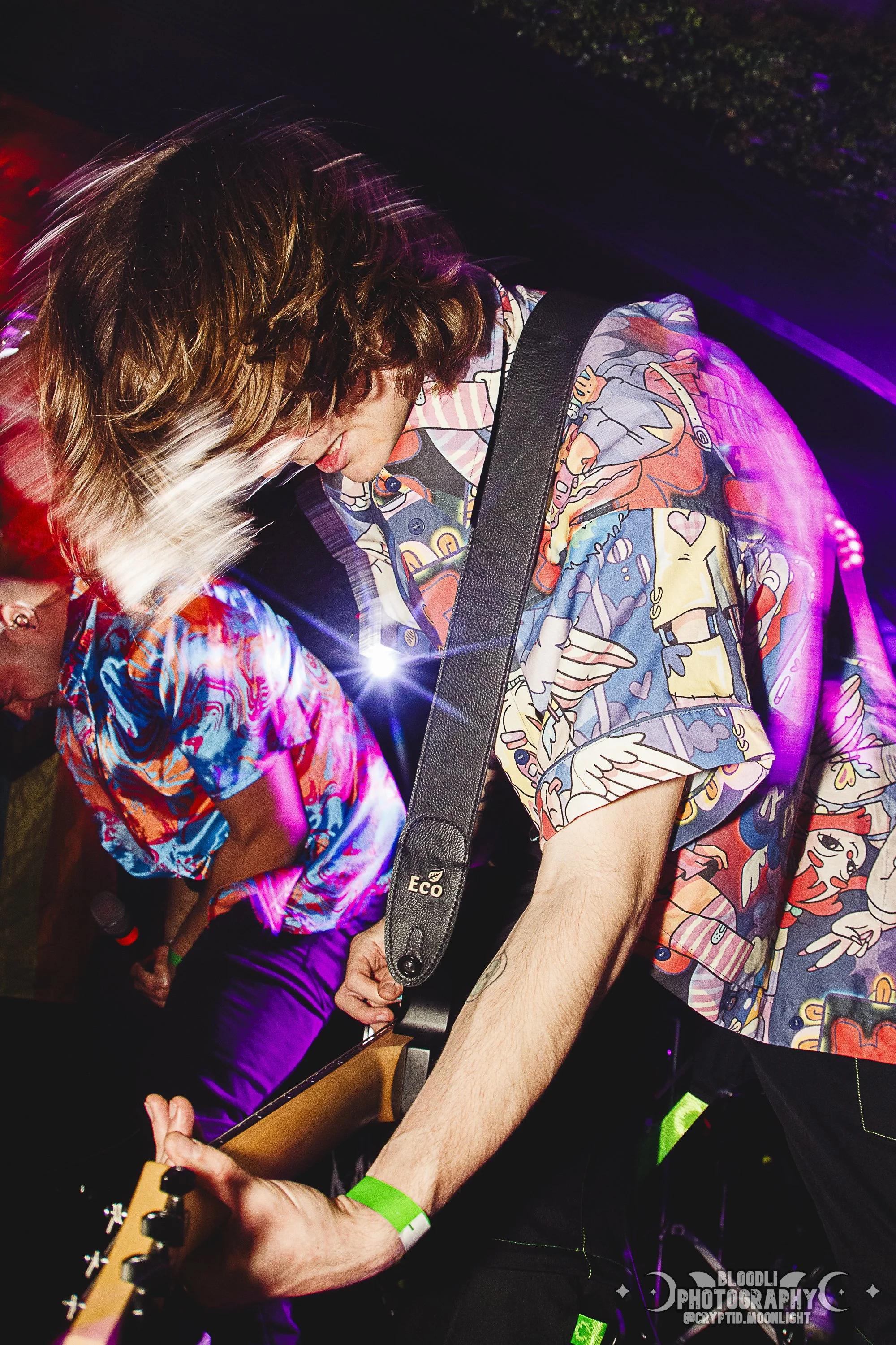 A man with wavy, shoulder-length hair wearing a colorful, patterned short-sleeve shirt playing an electric guitar at a lively event with purple and pink lighting.
