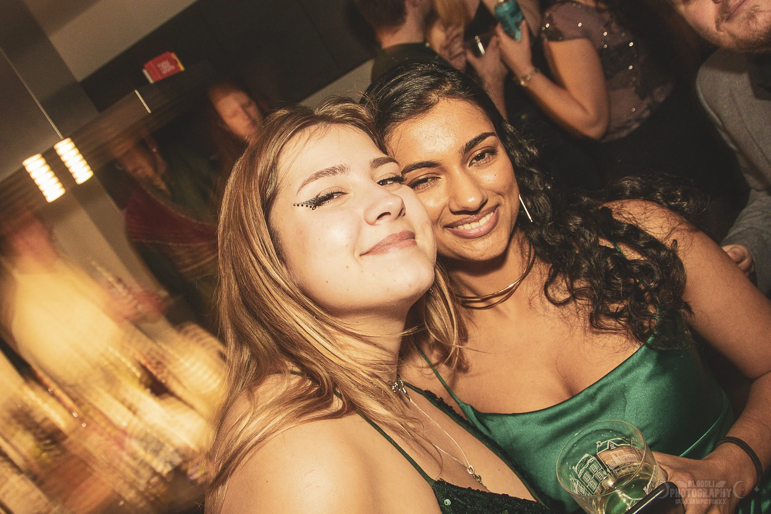 Two women smiling and posing for a photo at a party, with other guests in the background.