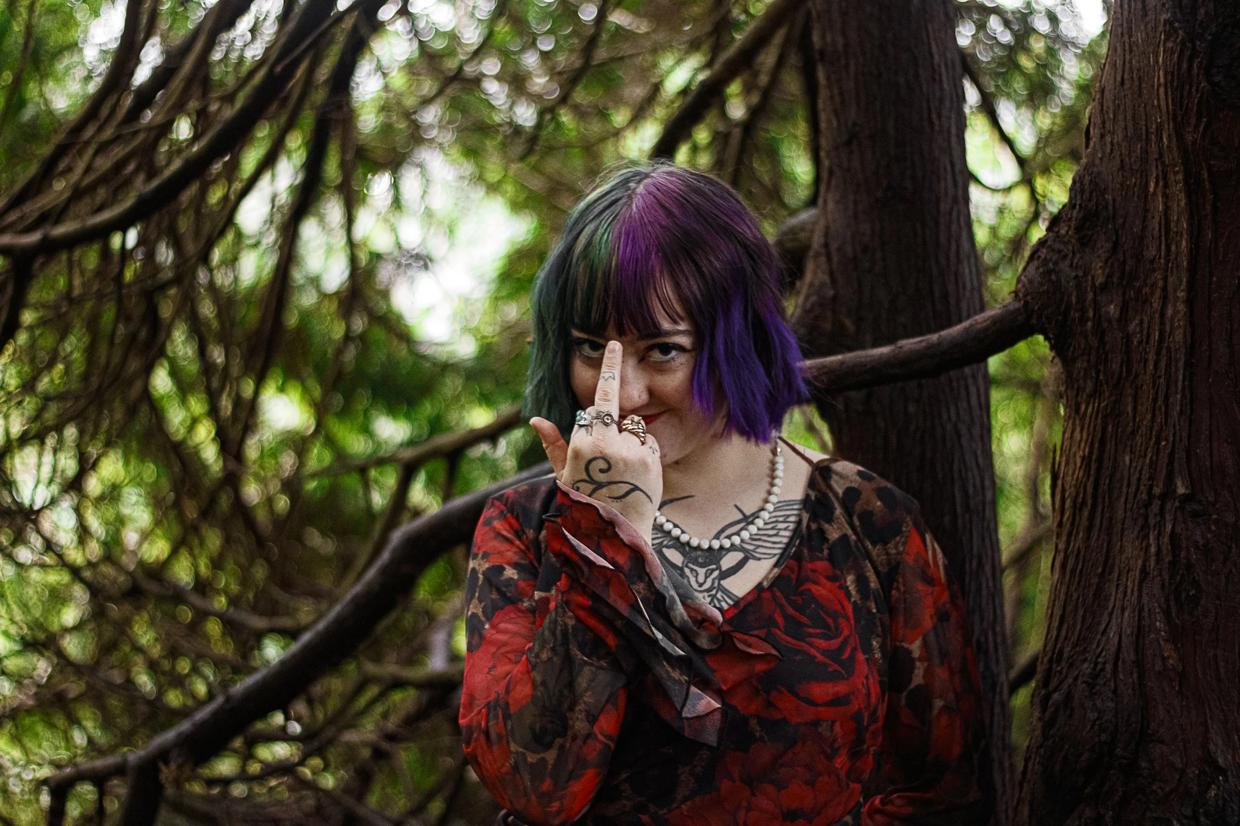 A woman with purple, green, and black hair, tattoos, and rings on her fingers, standing among trees with dense foliage. She is wearing a red and black patterned dress, a pearl necklace, and has her middle finger raised to her face, pointing at her no