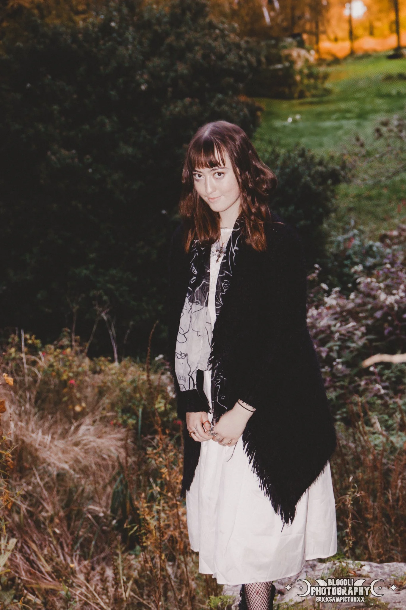 A young woman with shoulder-length brown hair and bangs standing outdoors in a garden with trees and flowers in the background, wearing a black fur coat, a patterned scarf, and a long white skirt.