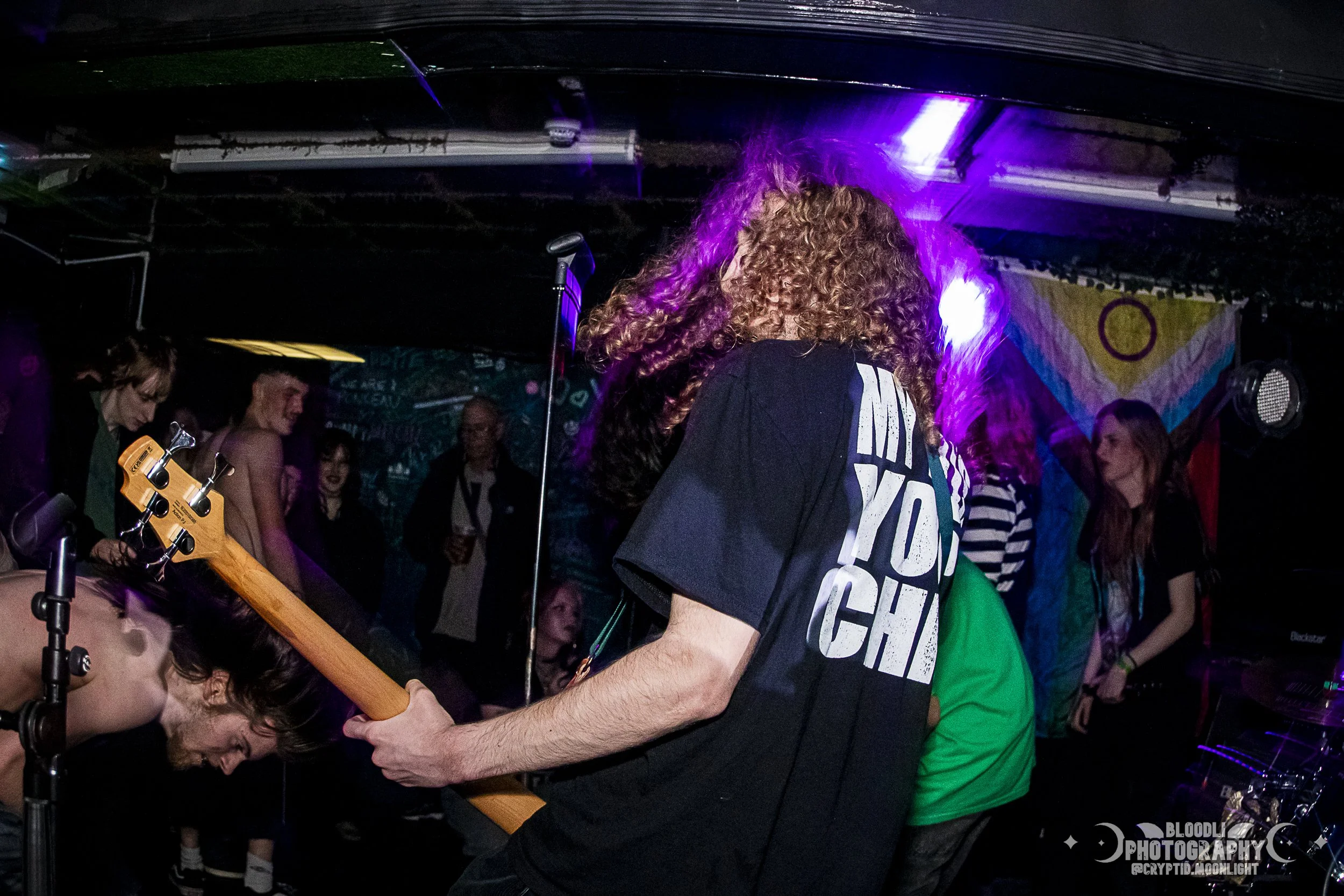 A musician with curly hair playing a bass guitar at a concert, with a colorful backdrop and audience in the background.