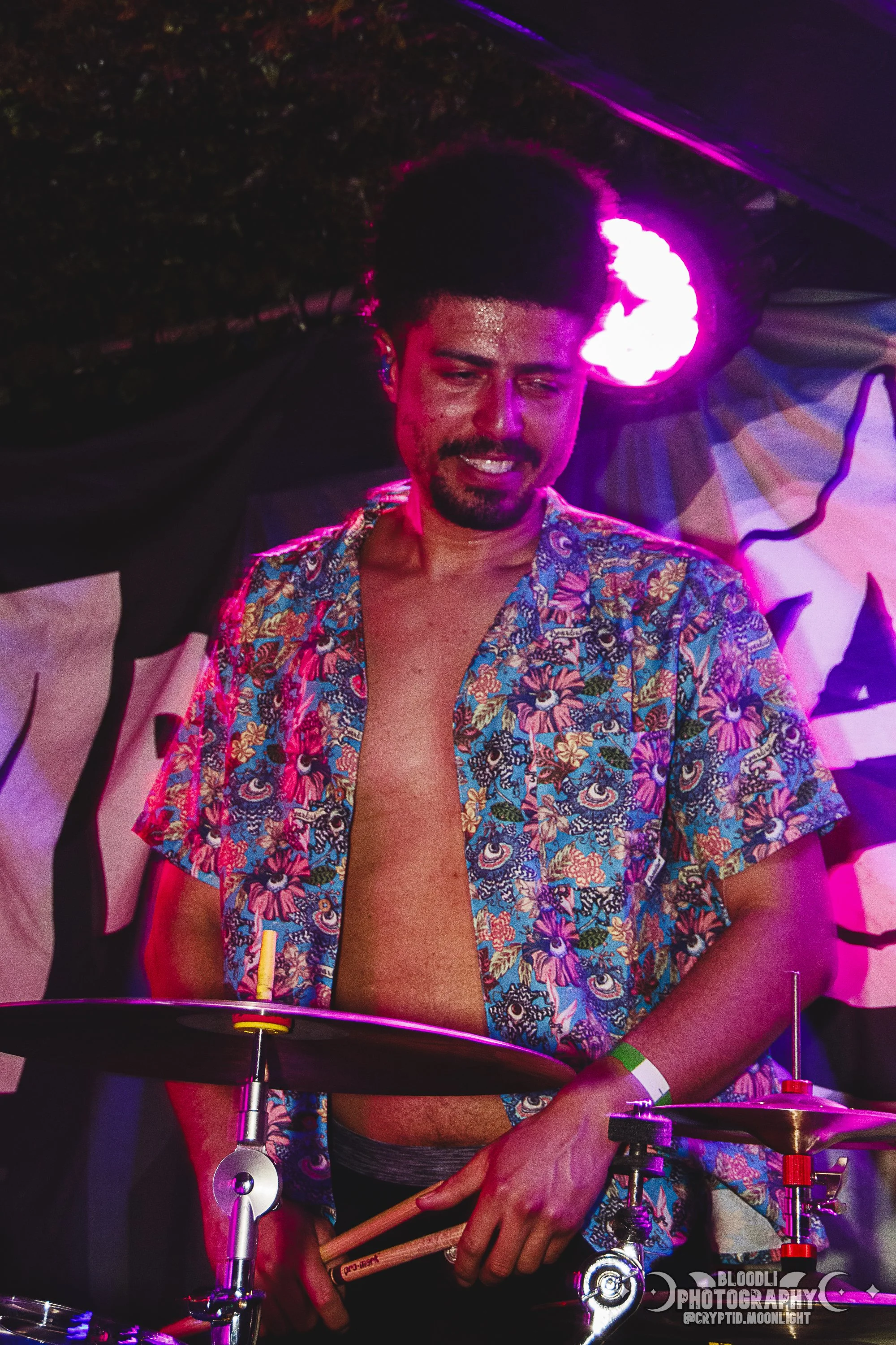 A man with dark curly hair, a beard, and a mustache standing behind a drum set, wearing a colorful floral Hawaiian shirt with no undershirt, under purple stage lighting during a performance.