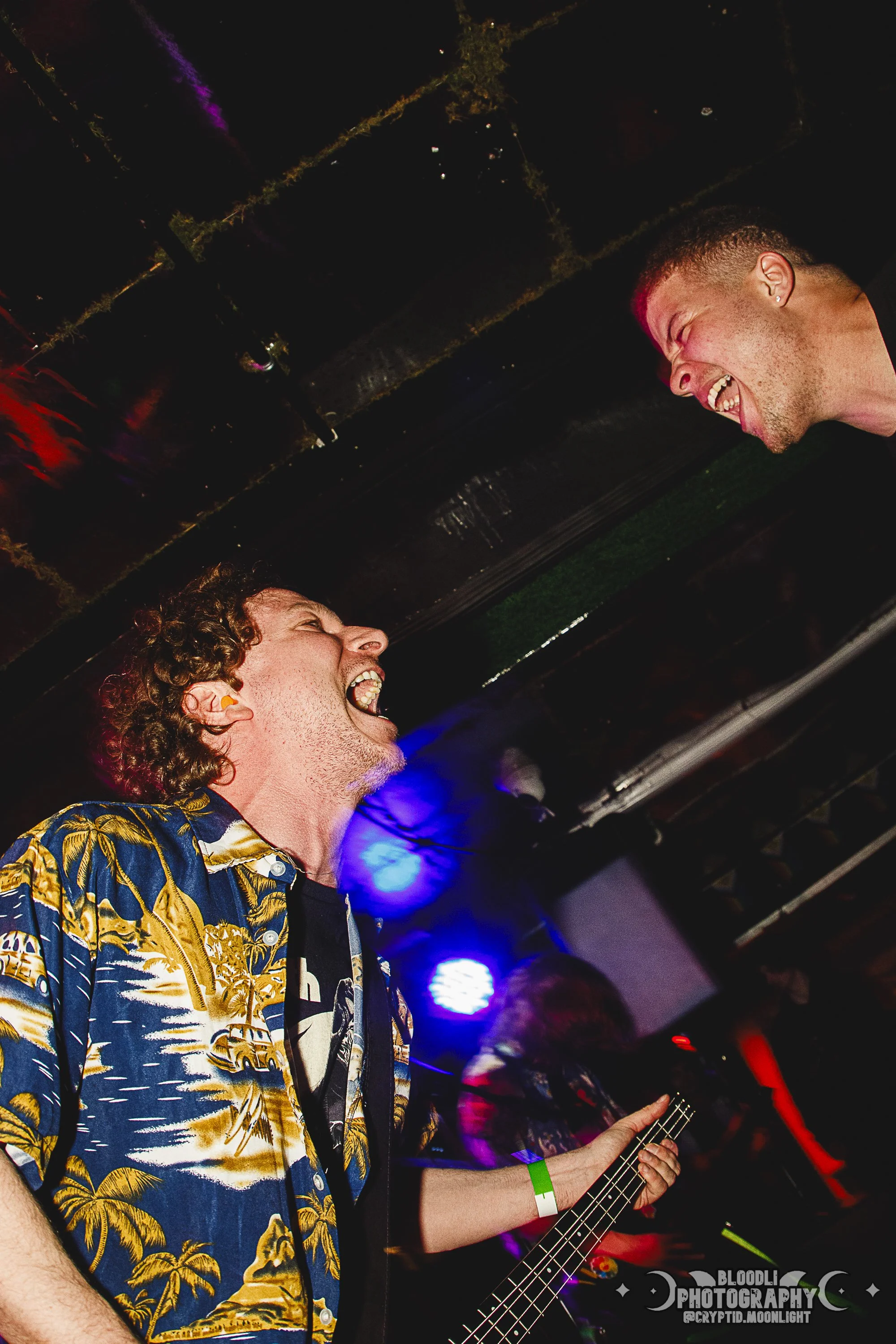 Two men are smiling and laughing at each other during a live music performance in a dimly lit venue with colorful stage lights, one holding a guitar.