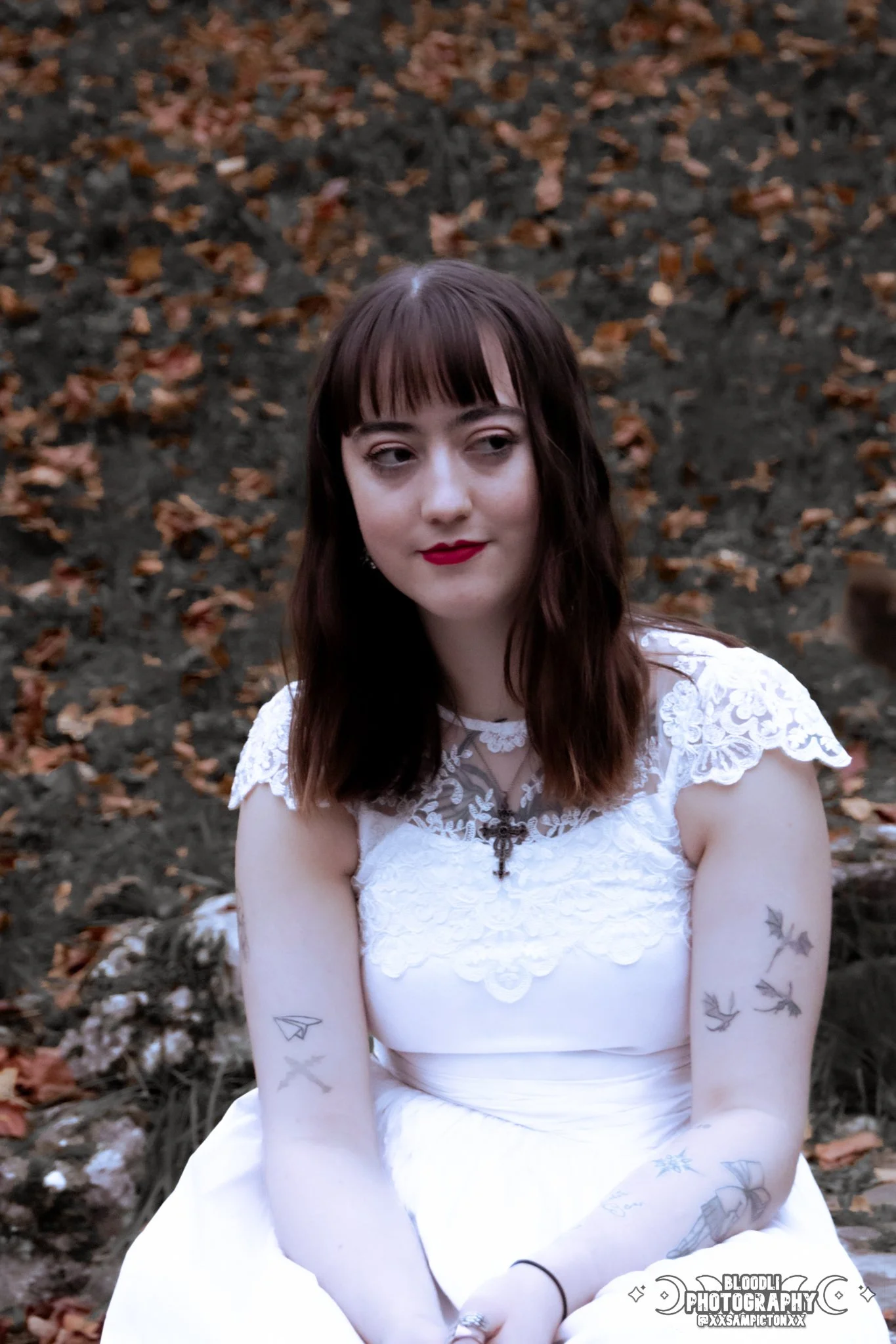 A young woman with dark brown hair wearing a white lace dress with tattoos on her arms, sitting outdoors on a rocky surface with fallen leaves in the background.