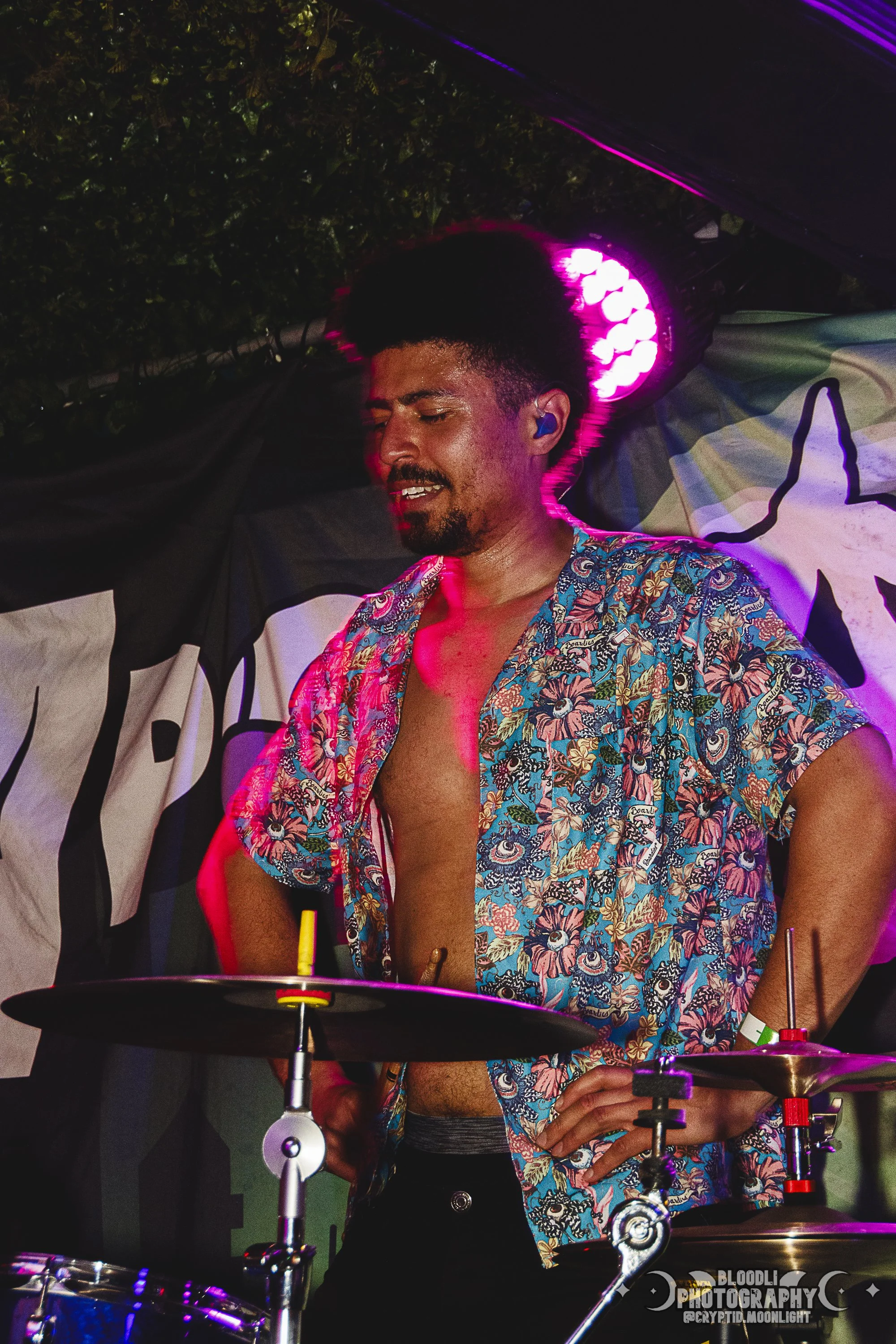 A man with curly black hair and a beard, wearing a colorful short-sleeved shirt partially open, playing drums during a performance at night with pink stage lighting.
