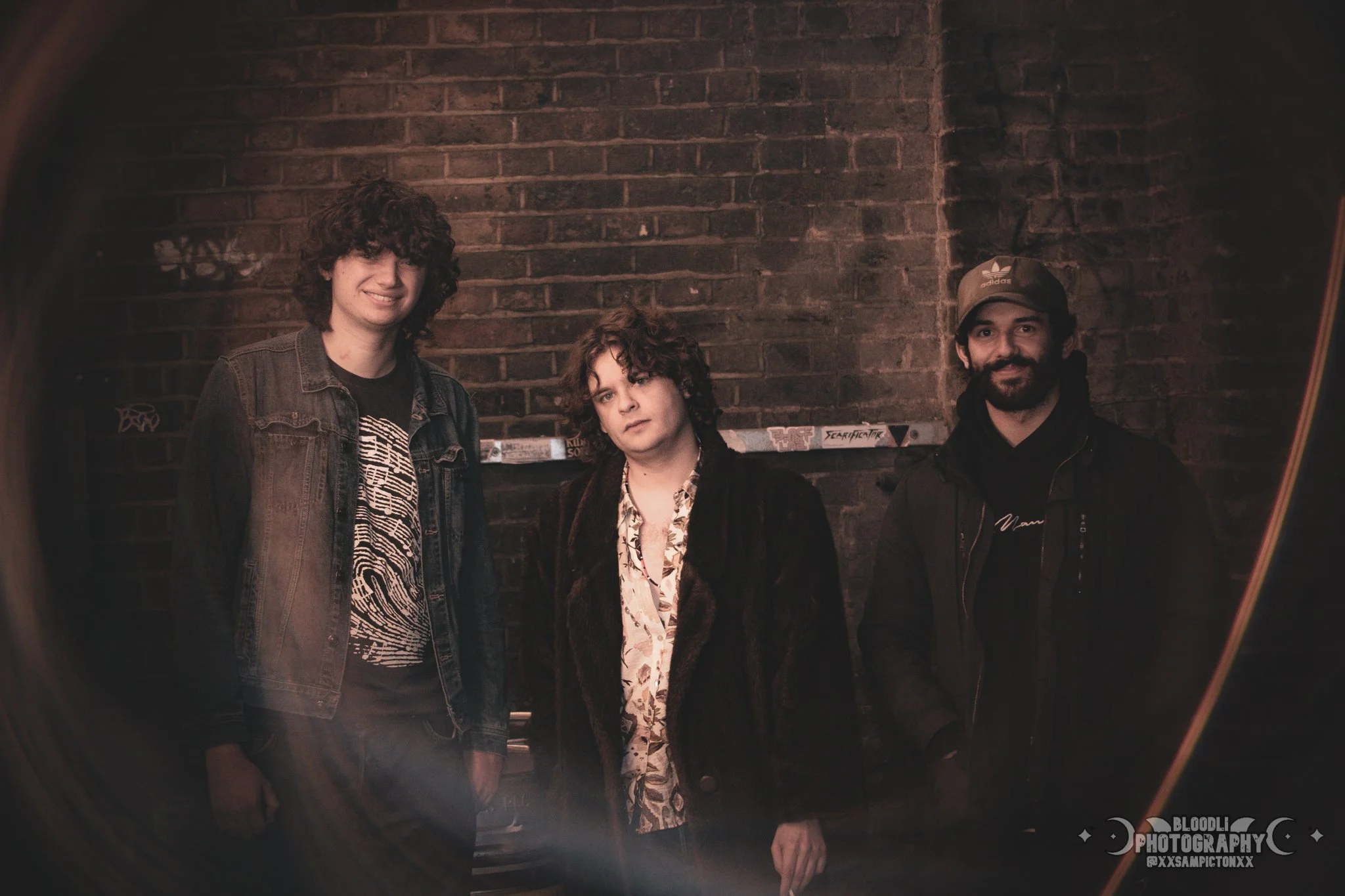 Three young men standing against a brick wall, looking at the camera, in a dimly lit setting with graffiti and stickers in the background.