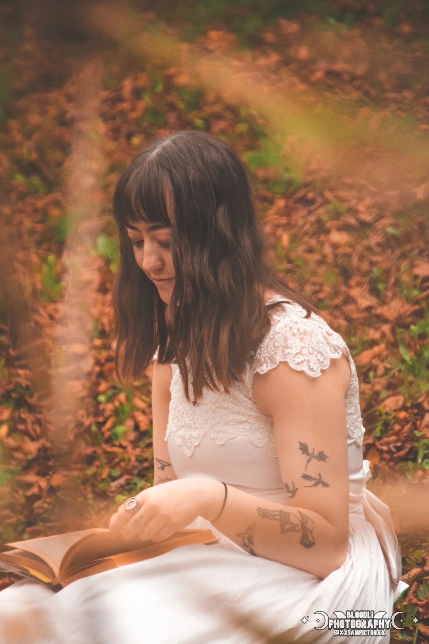 A woman with dark brown wavy hair and tattoos on her arm, sitting outdoors on autumn leaves, reading a book.
