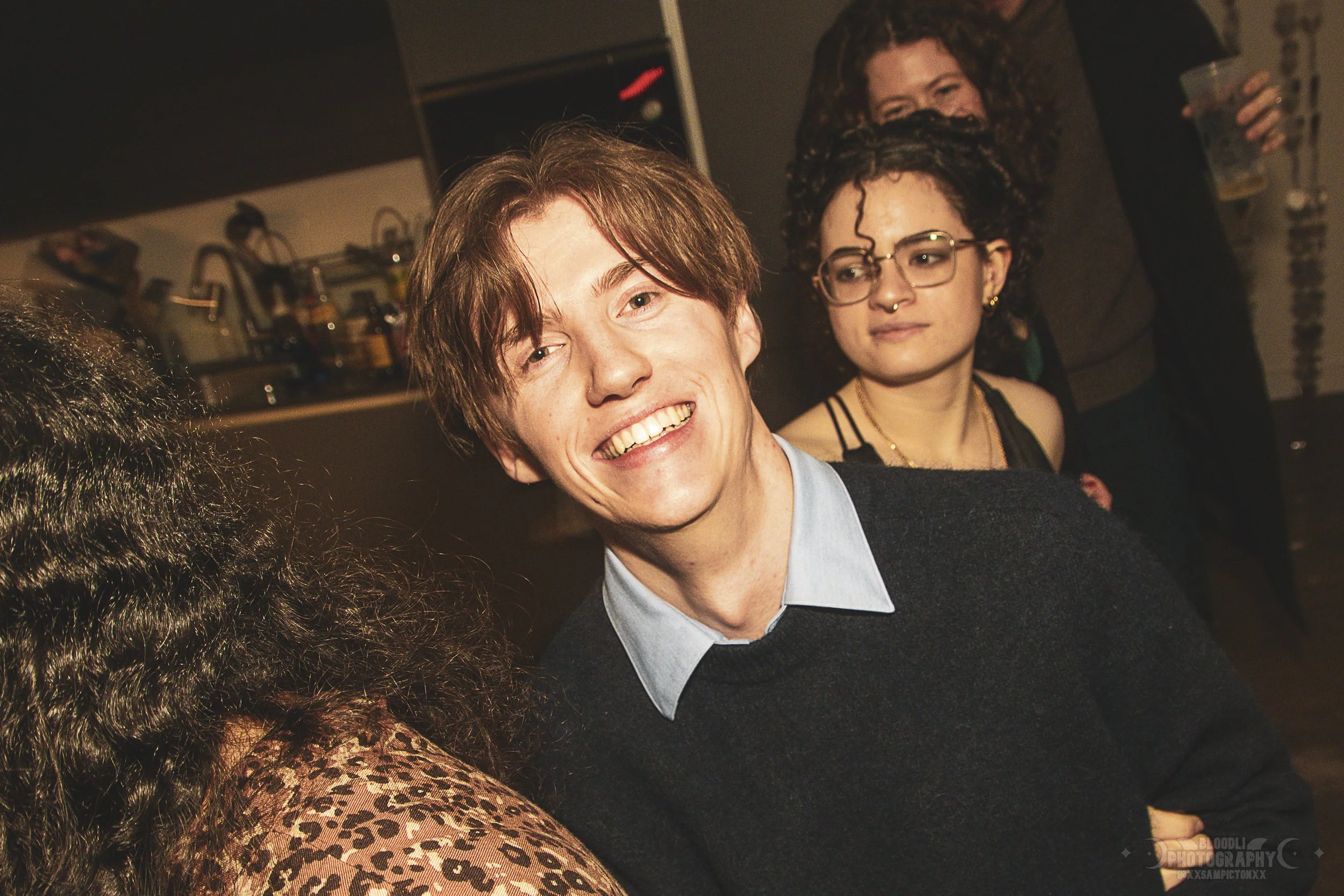 A young man with brown hair smiling and wearing a light blue shirt with a dark sweater, sitting at a social event with other people in the background.