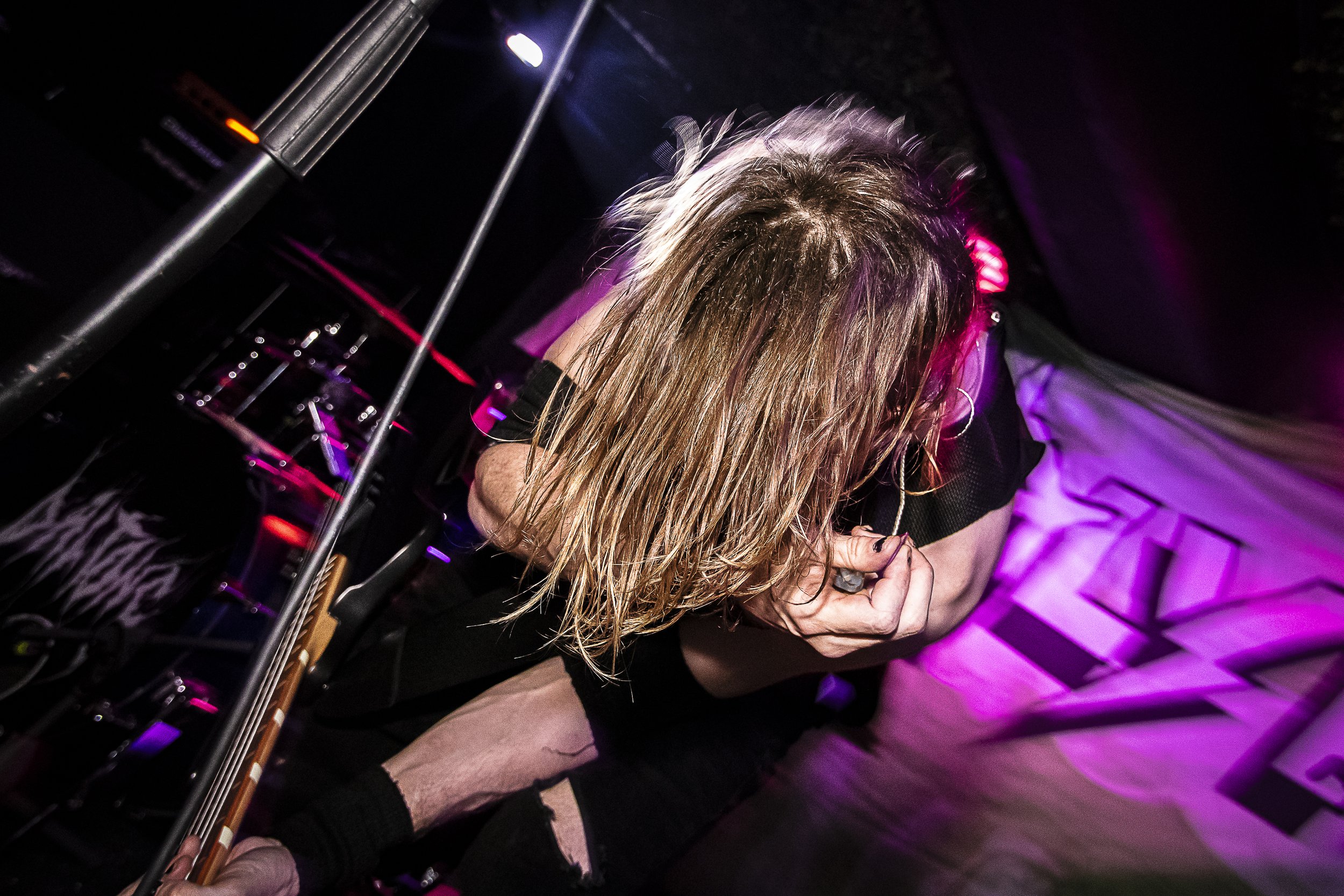A woman with long blonde hair, wearing a black top and ripped shorts, kneeling on stage with her head bowed and hand clenched into a fist.