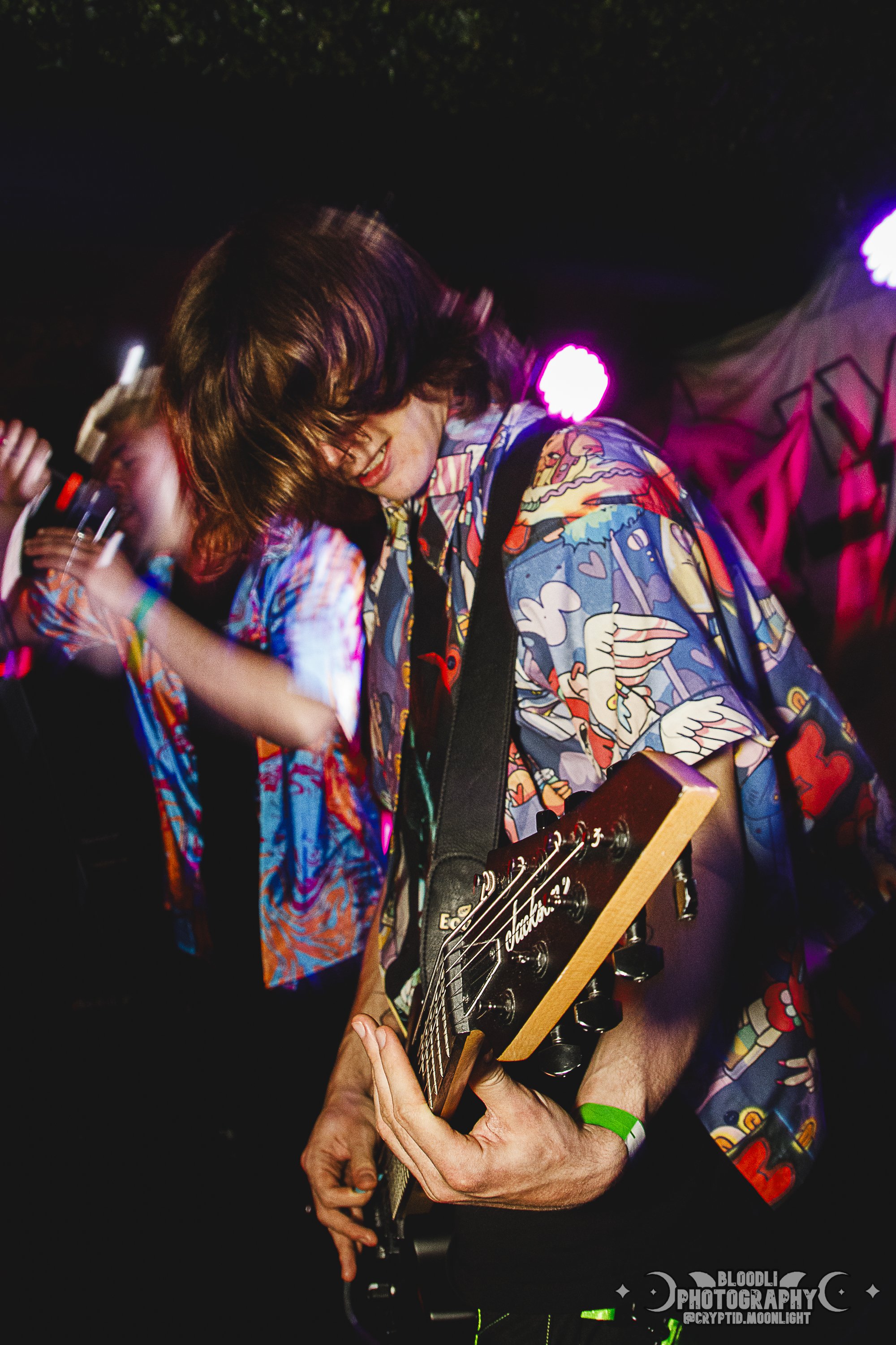 A young man playing an electric guitar on stage with colorful lighting, wearing a vibrant, patterned shirt.