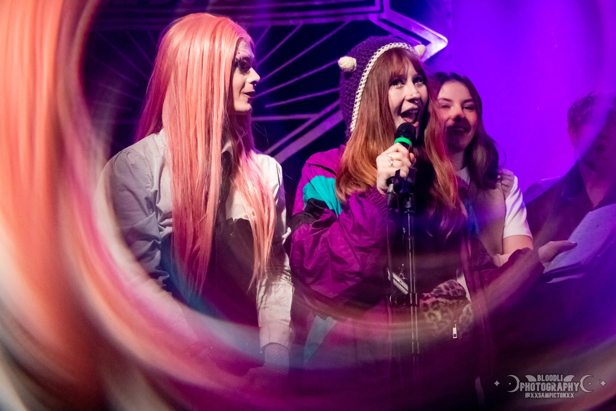 Group of people, including a woman wearing a purple jacket and a knit hat with bear ears, speaking into a microphone during a performance or event, with colorful purple and pink lighting in the background.