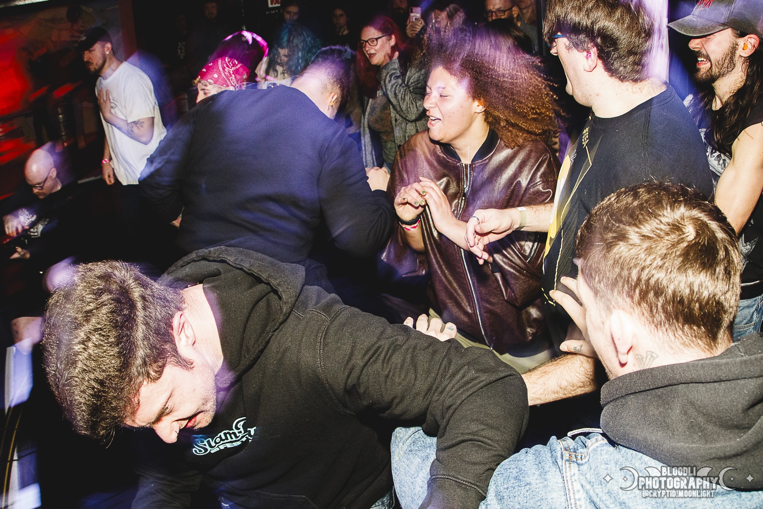 A crowd of people having a lively time at a concert or nightclub with colorful lighting, some dancing and others engaging in an energetic dance fight or wrestling match.