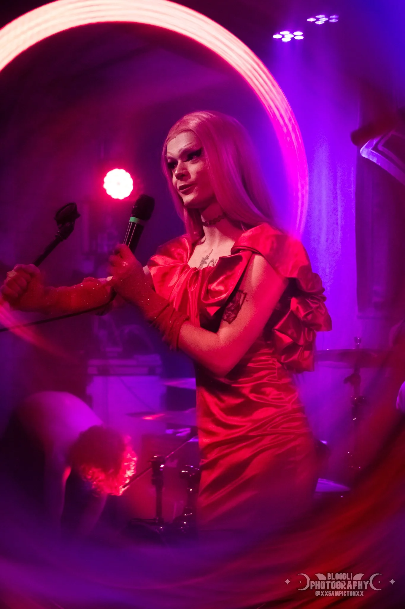 A person with long blonde hair performing on stage, holding a microphone, wearing a shiny red dress, with colorful stage lighting creating a vibrant atmosphere.