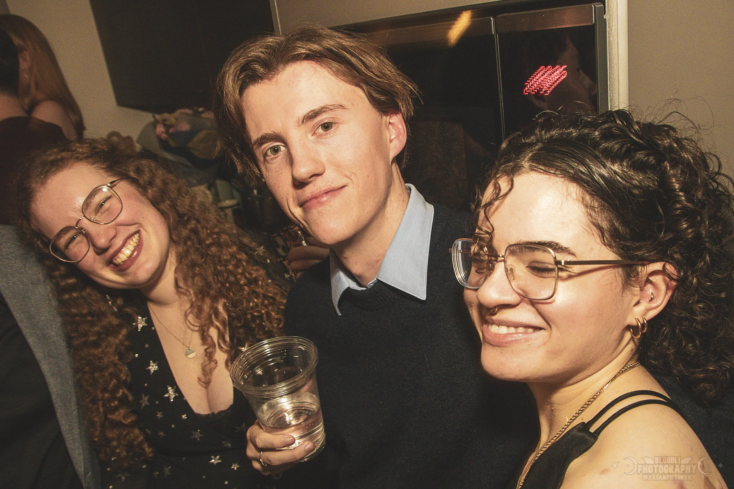 Three young people smiling and posing for a photo at a party, with one holding a cup of water.