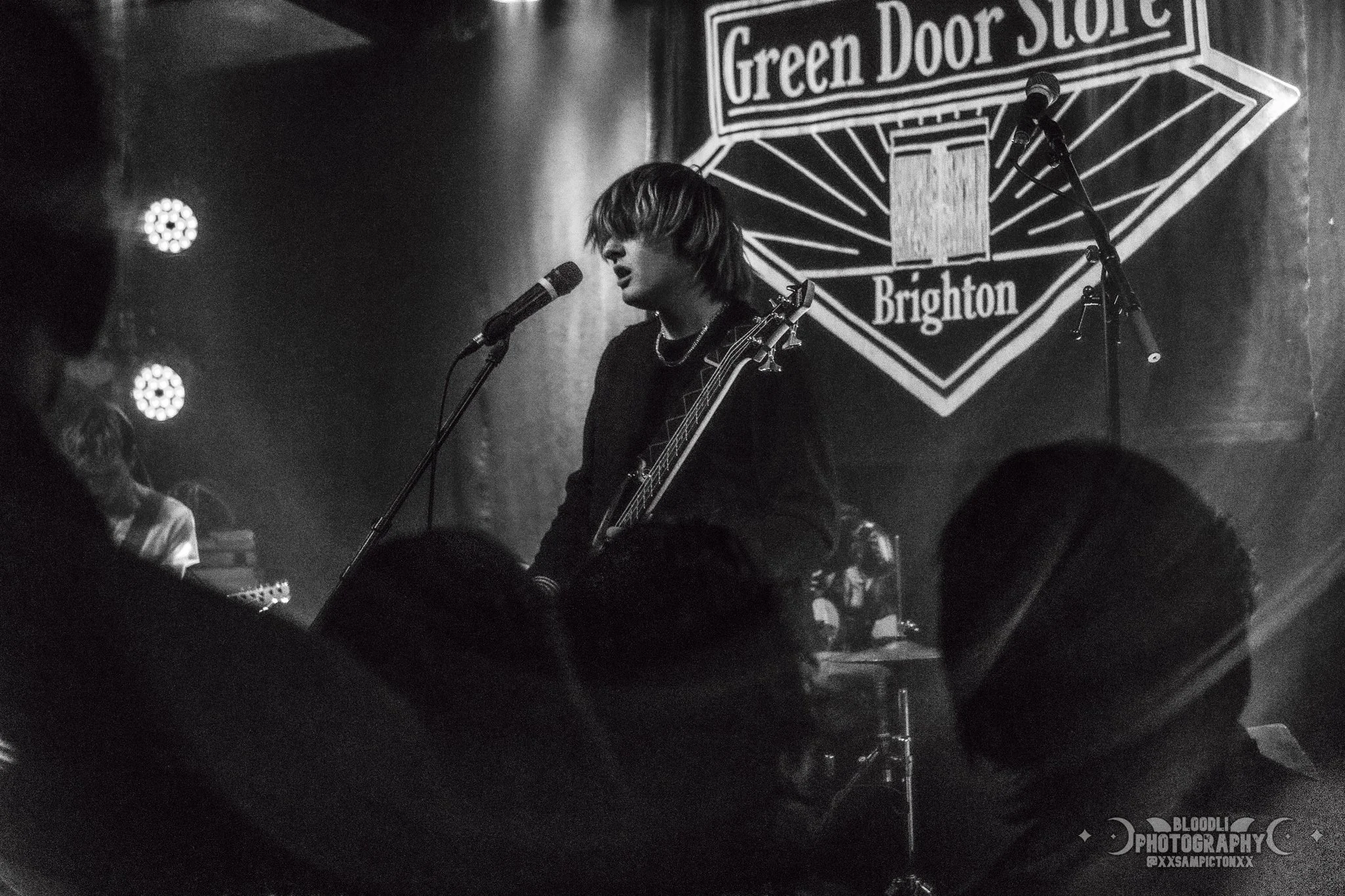 A musician performing on stage at Green Door Store Brighton, singing into a microphone and playing an electric guitar, with an audience in the foreground.