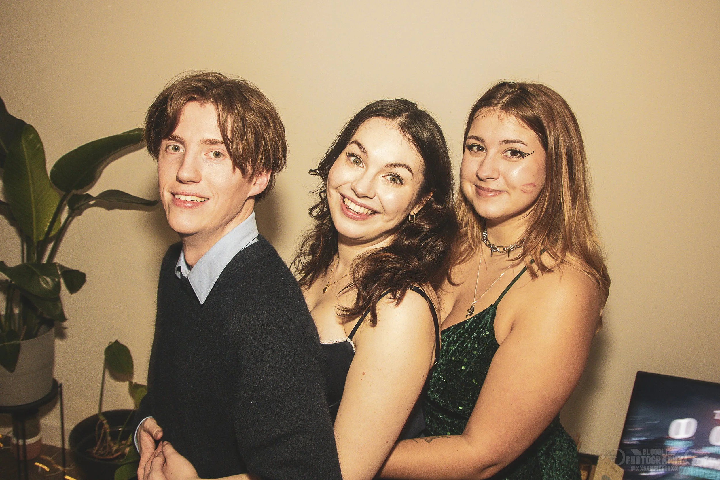 Three young adults smiling and posing together indoors, standing close to each other, with a potted plant on the left and a computer screen partially visible on the right in the background.