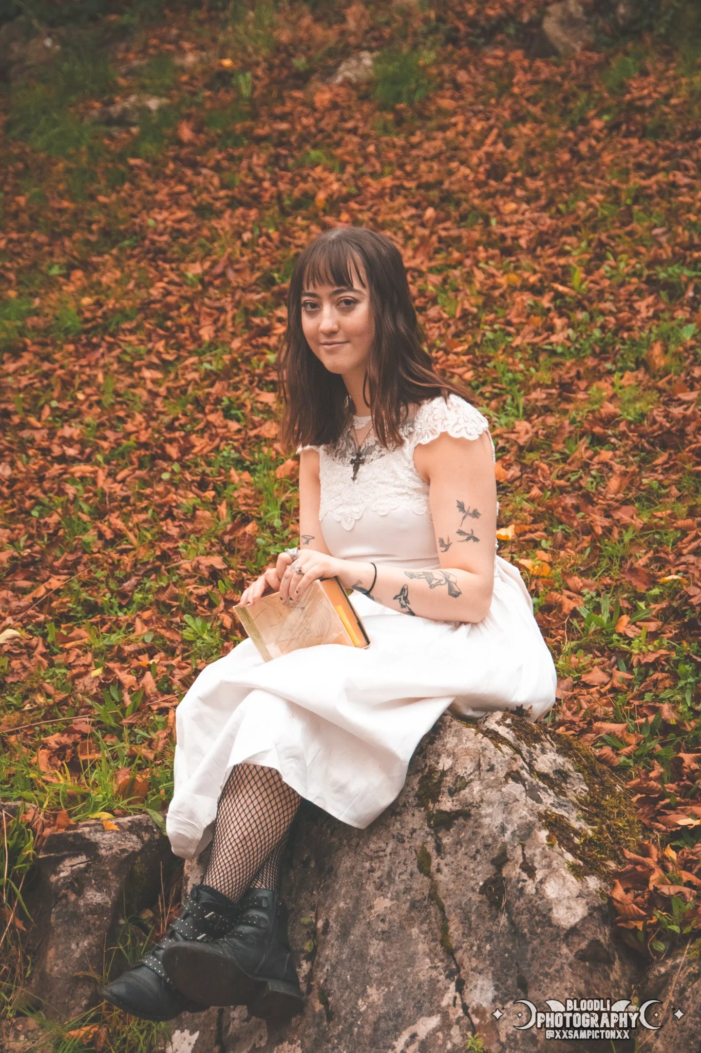 A young woman with dark brown hair and tattoos on her arms is sitting on a rock in a forested area covered with fallen leaves. She is wearing a white, lace-detailed dress, black fishnet stockings, and black combat boots. She is holding an open book i