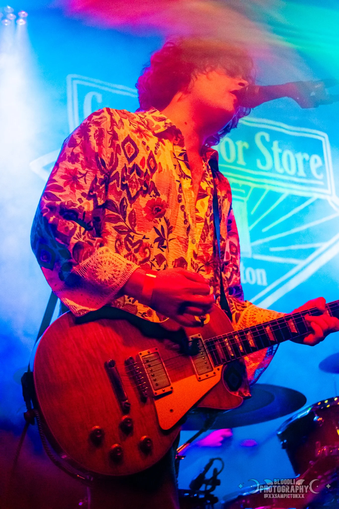 Musician playing electric guitar on stage with colorful lighting and a background sign that reads 'Guitar Store'.