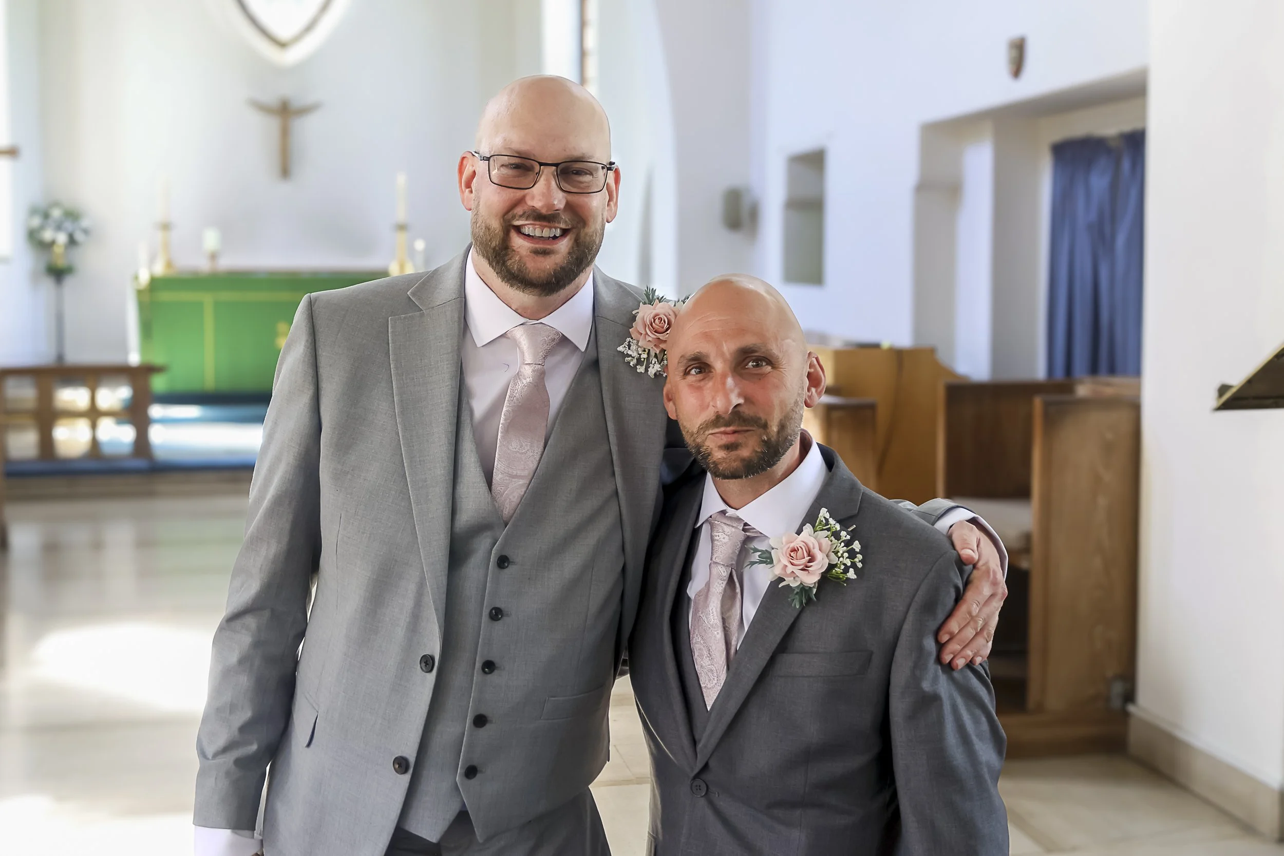 Two men in suits, one taller with glasses and a beard, and one shorter with a shaved head and beard, smiling and standing inside a church with religious symbols in the background.