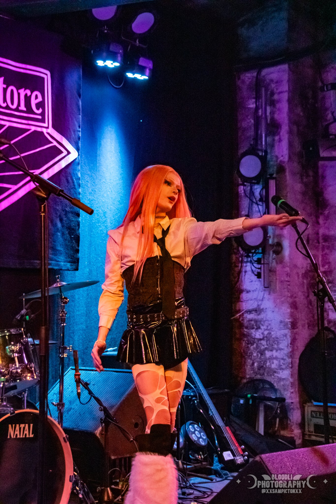 A performer with long orange hair singing on stage in a punk rock outfit, pointing towards the audience with her right hand, with stage lights and musical equipment around her.